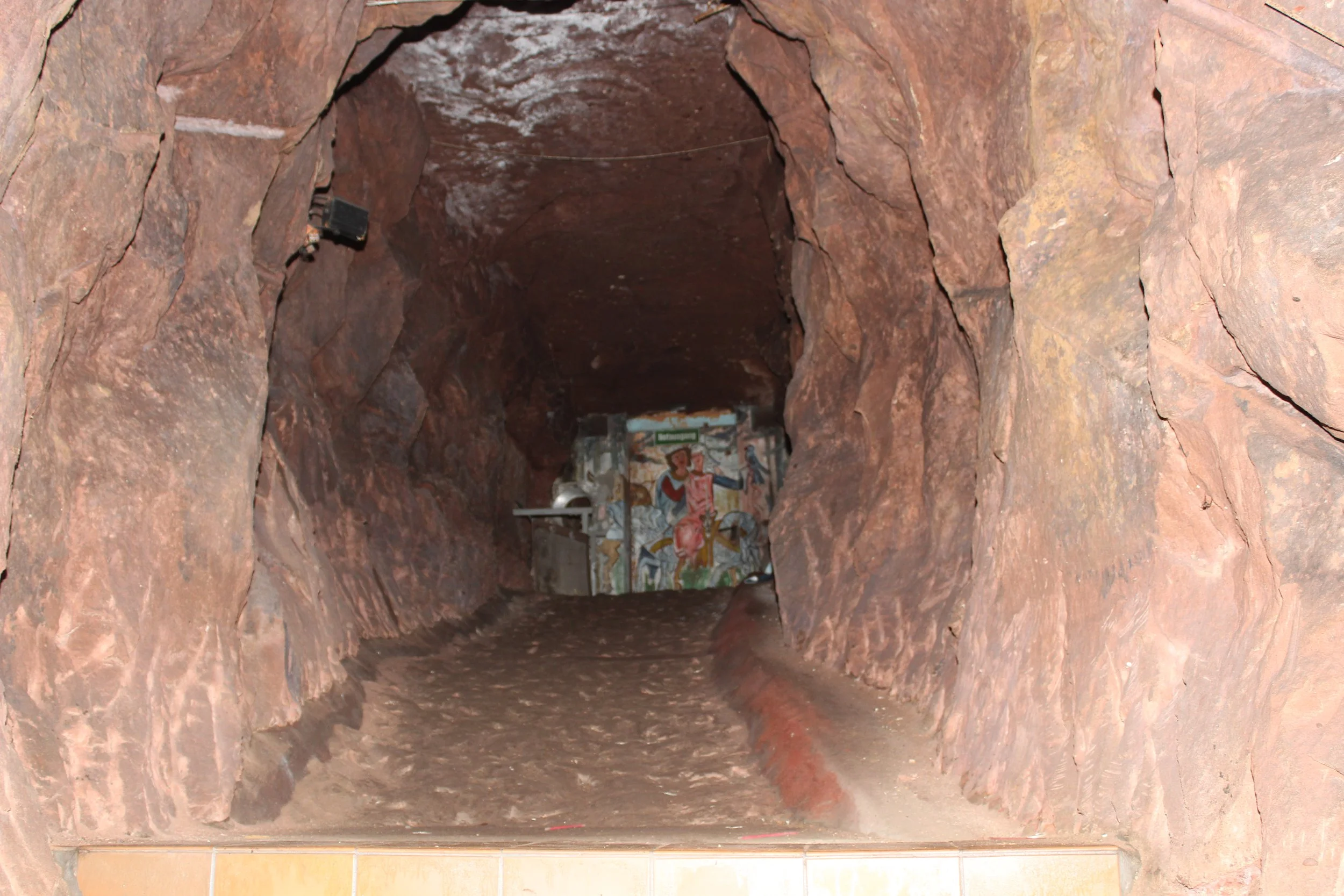 Ein erdachter Tunnel mit rotem Fels, im Hintergrund eine Wand mit bemalter Figur und einigen Gegenständen.