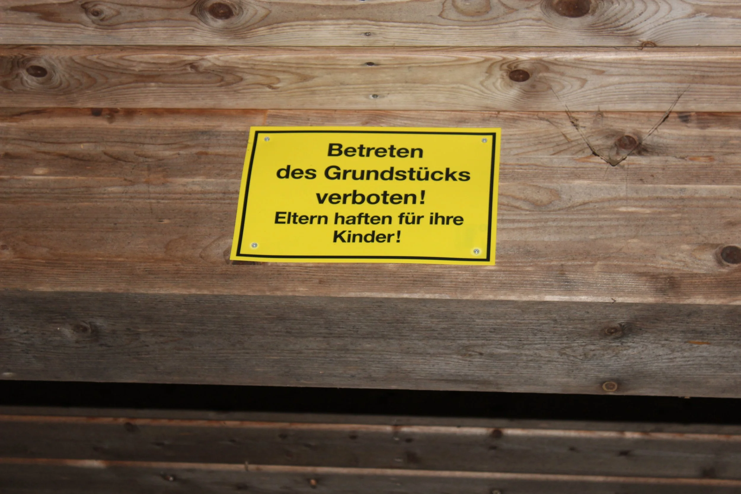 Gelbes Schild mit schwarzen Texten, das auf einer Holzfläche angebracht ist und das Betreten eines Grundstücks verbietet, wobei Eltern für ihre Kinder haften.