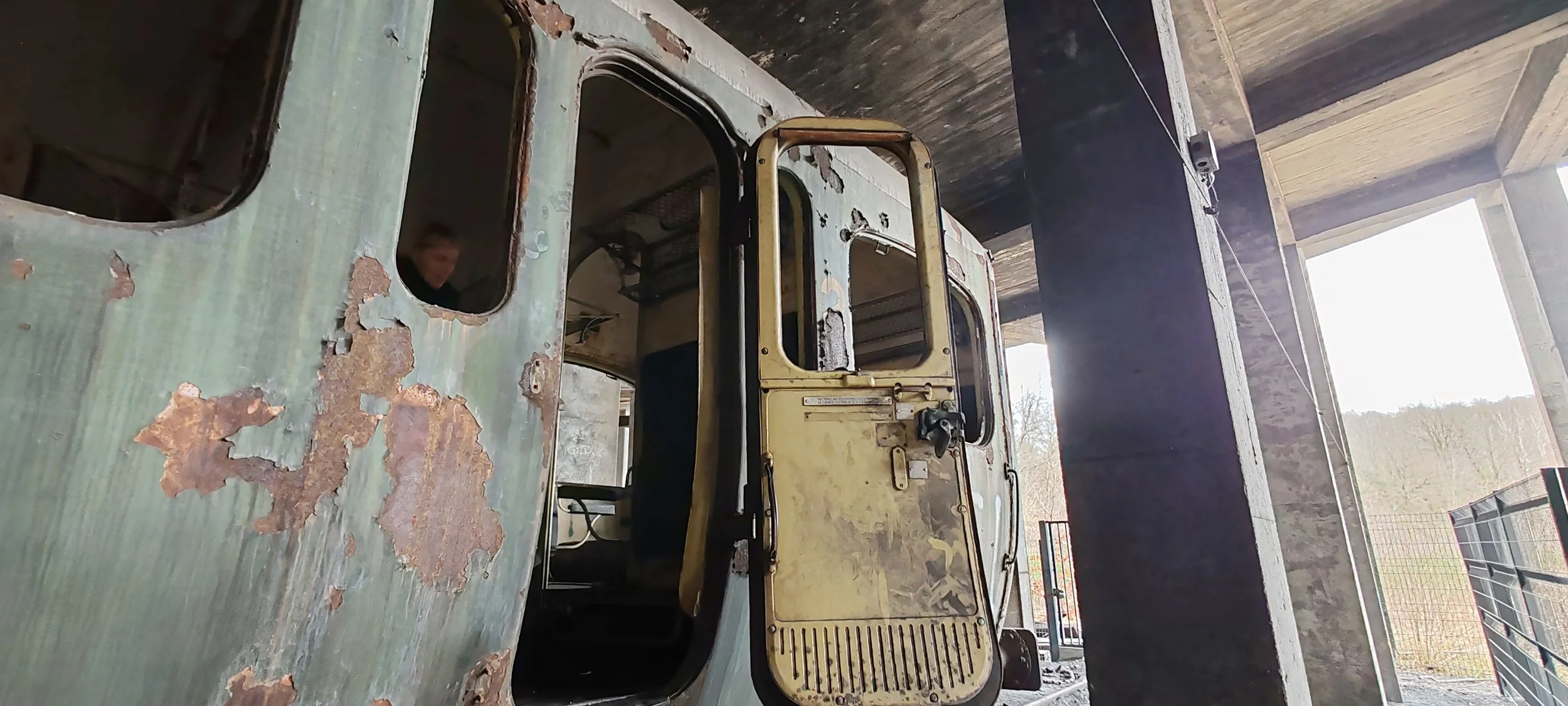 Verrosteter alte Bus in einem verfallenen Gebäude mit offener Tür, im Hintergrund offene Landschaft und Bäume sichtbar.