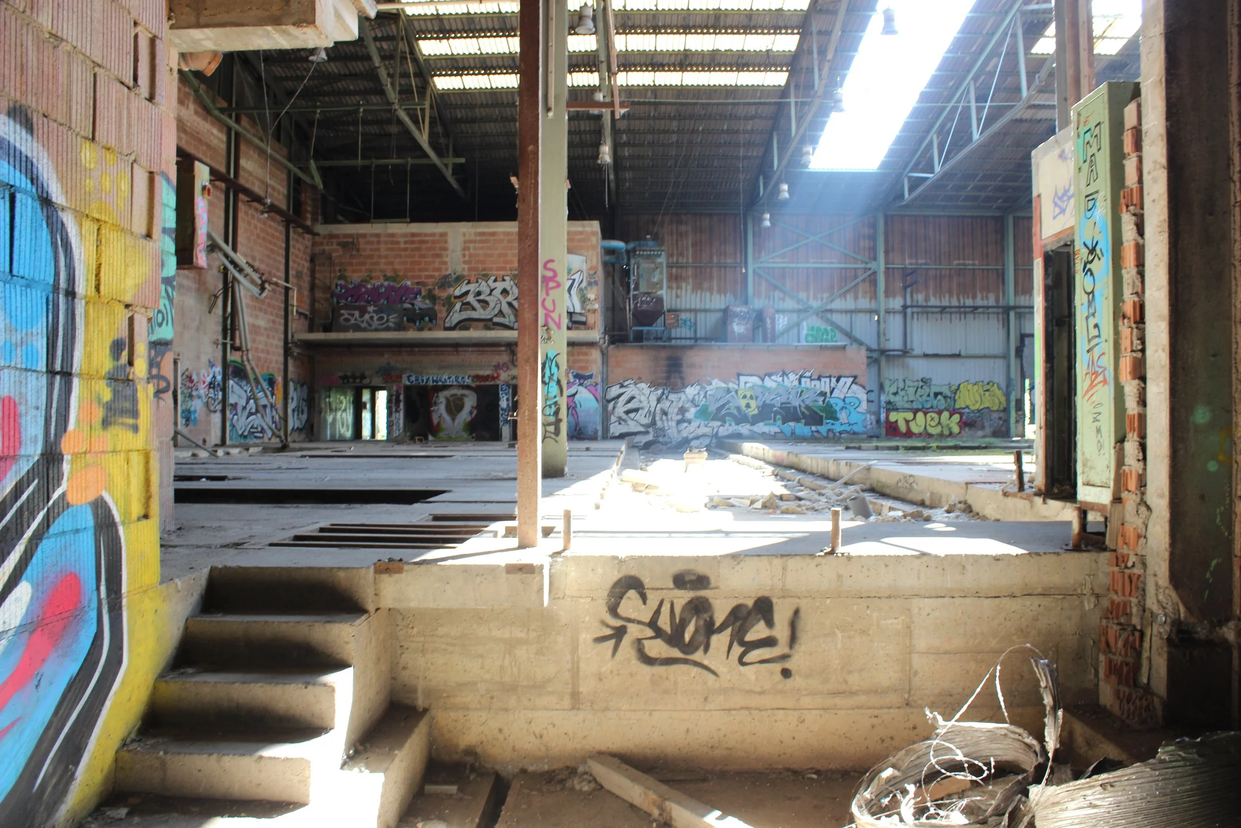 Innenansicht eines verlassenen Gebäudes mit Graffiti an den Wänden, teilweise zerstörtem Boden und Sonneinschienen von oben.