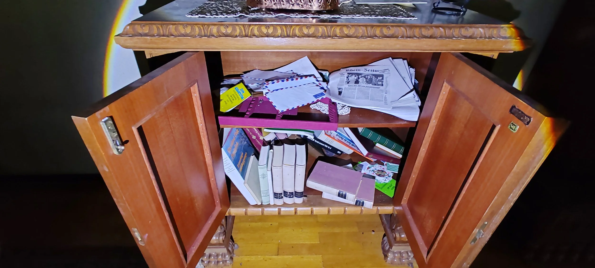 Ein aufgeräumtes Holzschränkchen mit zwei offenen Türen, gefüllt mit Büchern, Papieren, Umschlägen, Karten und Schreibutensilien, steht auf einer Holzbohle.