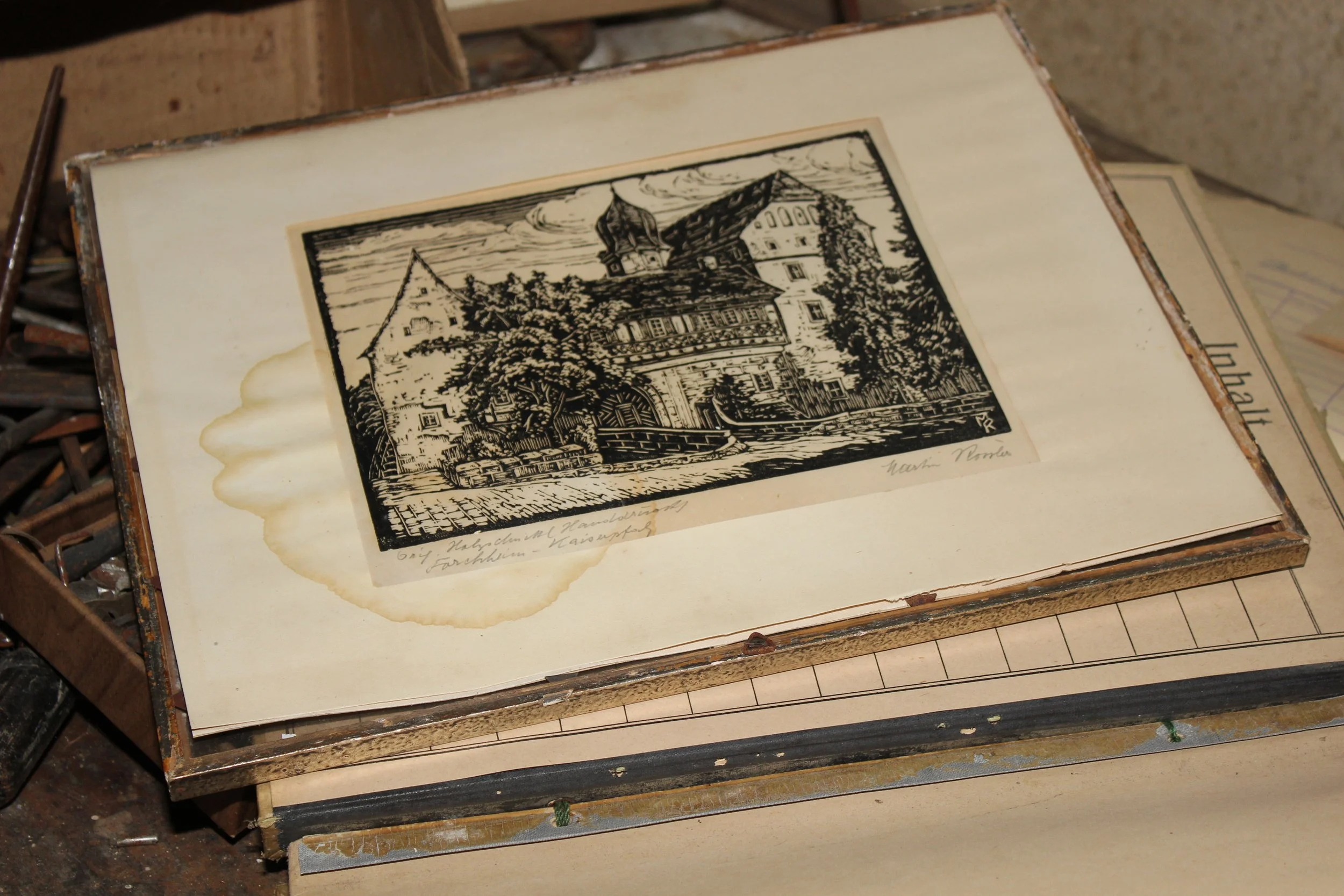 Ein altes Schwarz-Weiß-Holzschnittbild eines Hauses mit Wasser und Bäumen, im Rahmen einer alten Mappe oder eines Albums.