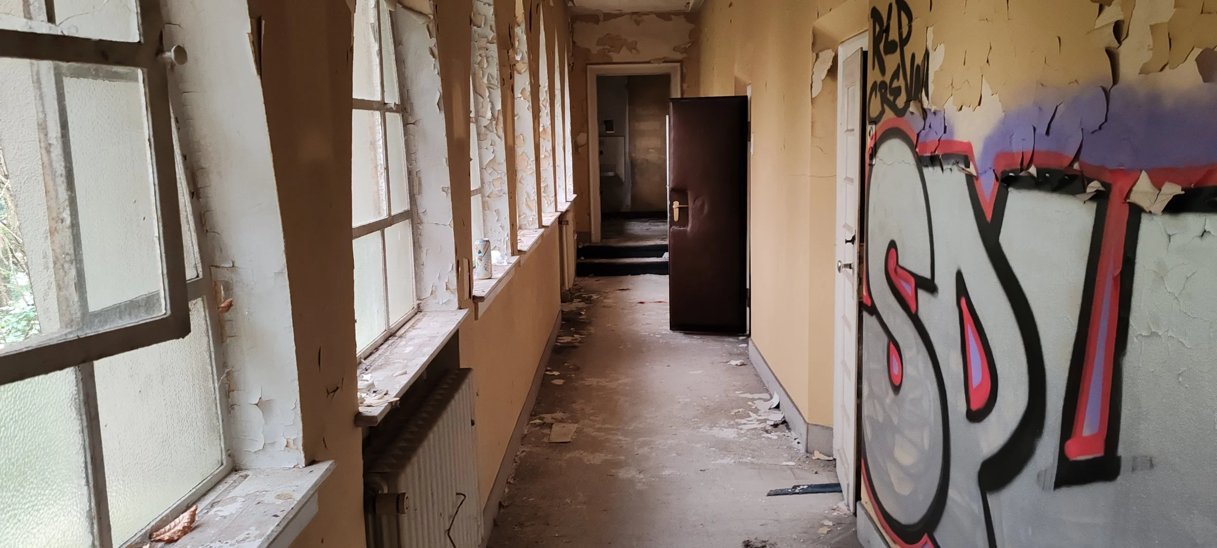 Ein alter, verlassener Flur mit zerbrochenen Fenstern, abblätternder Wandfarbe und Graffiti an der Wand.
