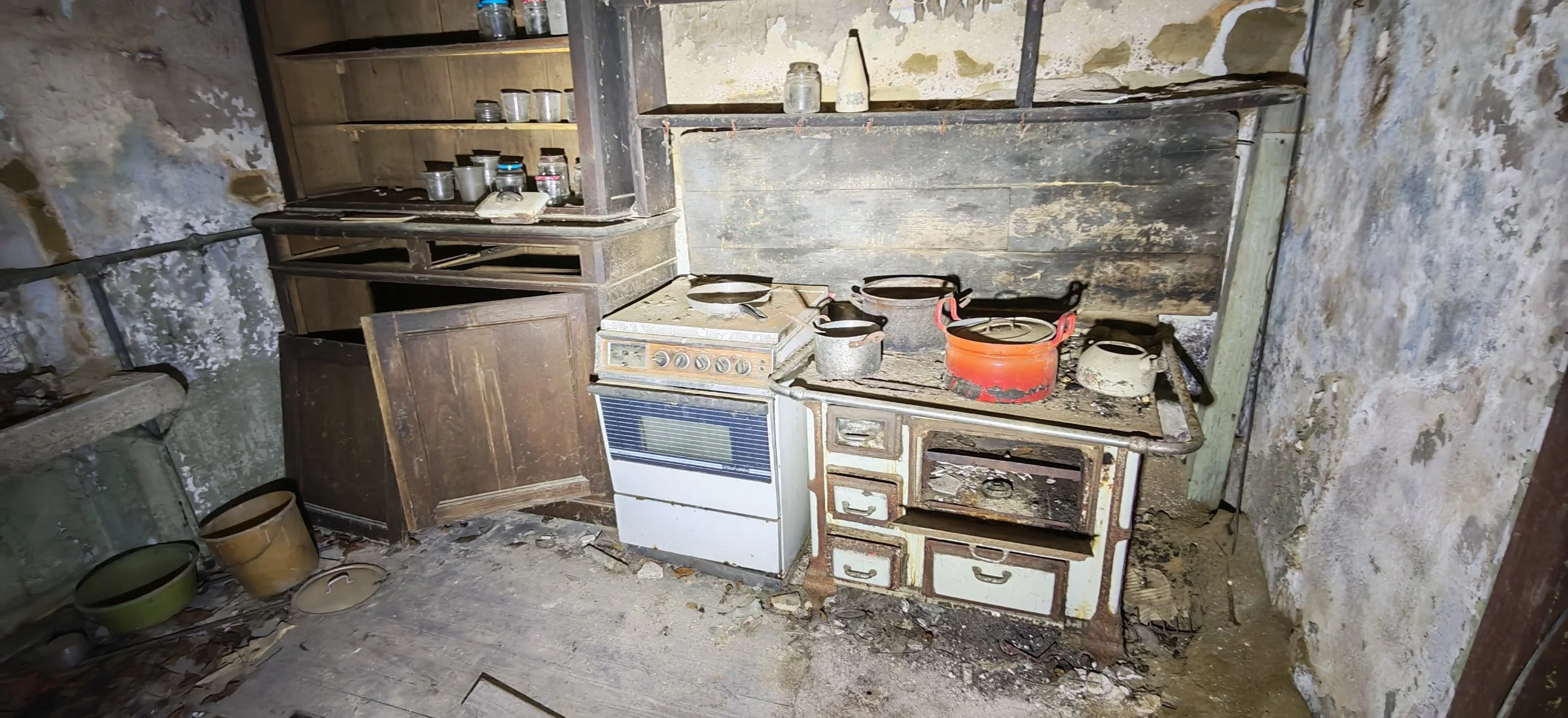 Verlassenes, verstaubtes Küchenkabinett mit alter, beschädigter Elektroherd, verrostetem Kamin, Staub und Schmutz auf dem Boden.