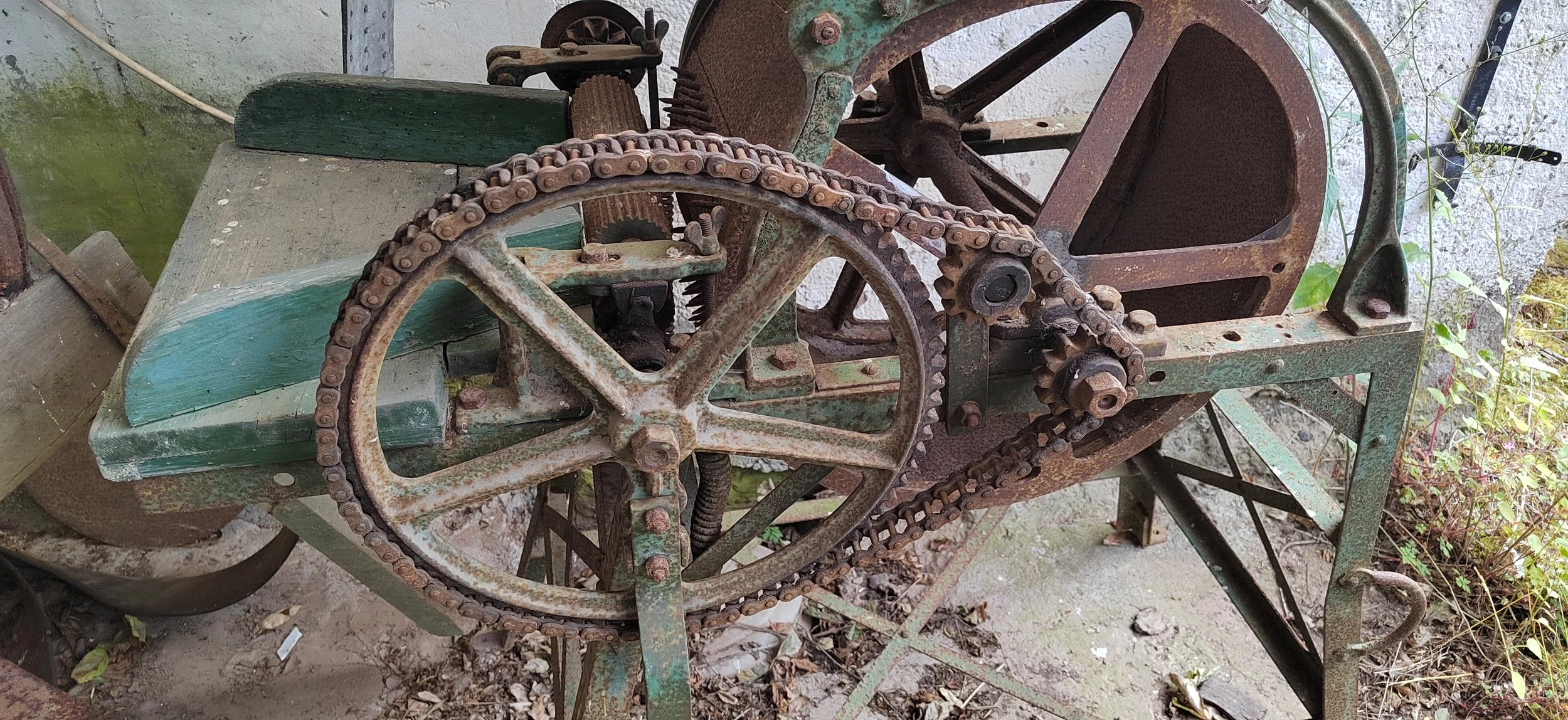 Verrostete alte Maschine, vermutlich eine antike Rührmaschine oder Schneidemaschine, mit großen Zahnrädern und Ketten, auf einer staubigen, verfallenen Außenfläche mit Pflanzen im Hintergrund.