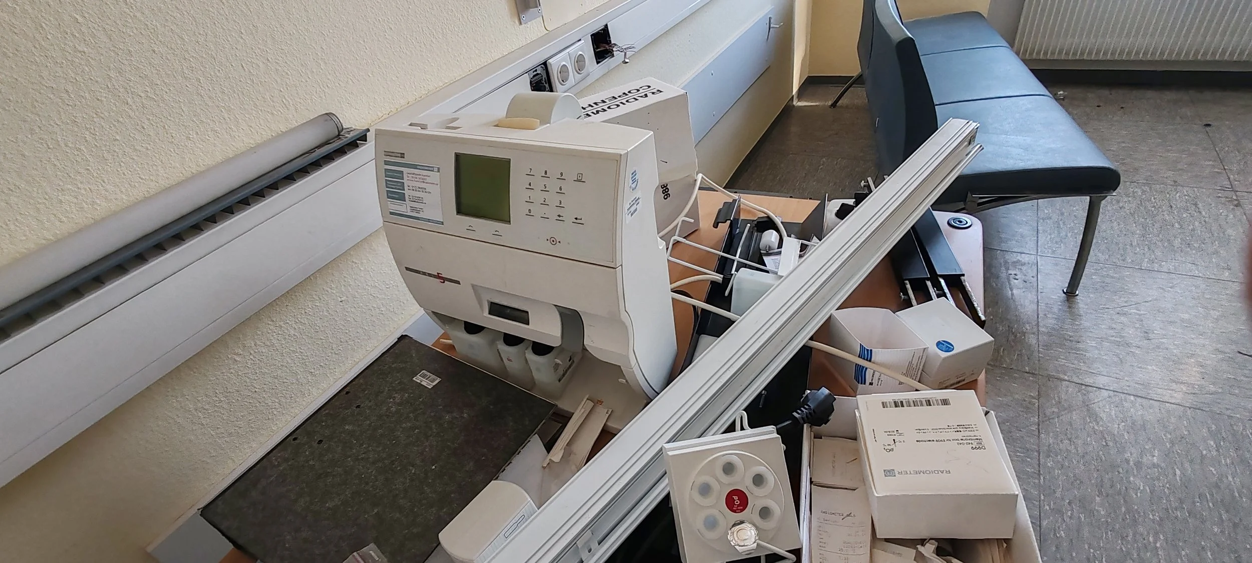 Ein Tisch mit medizinischer Ausrüstung, einschließlich eines Blutdruckmessgeräts, verschiedenen Verpackungen, Kabeln und einem Stift. Im Hintergrund eine Bank und eine Heizungsrohr an der Wand.