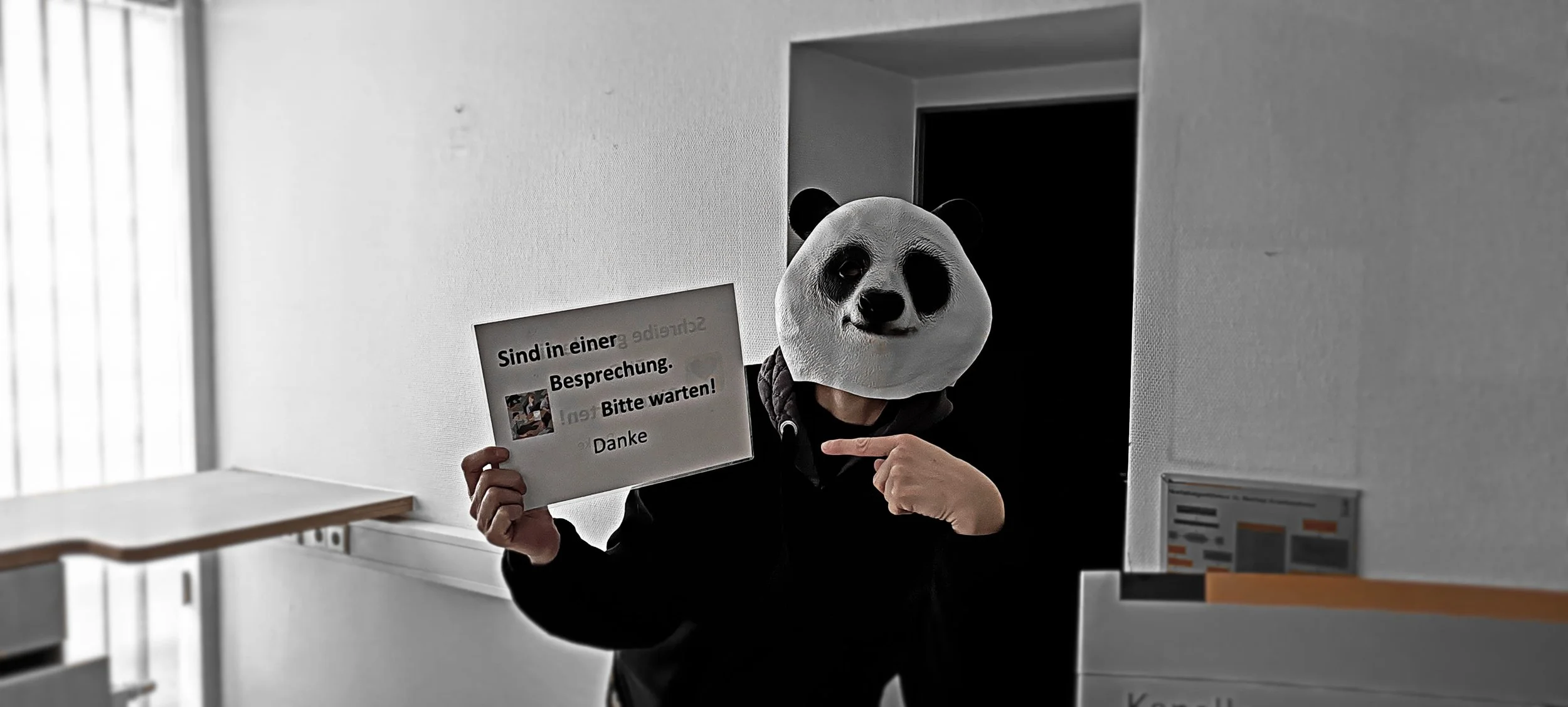 Person mit Pandamaske hält Schild mit Hinweisen, in einem Raum, vermutlich an einer Rezeption