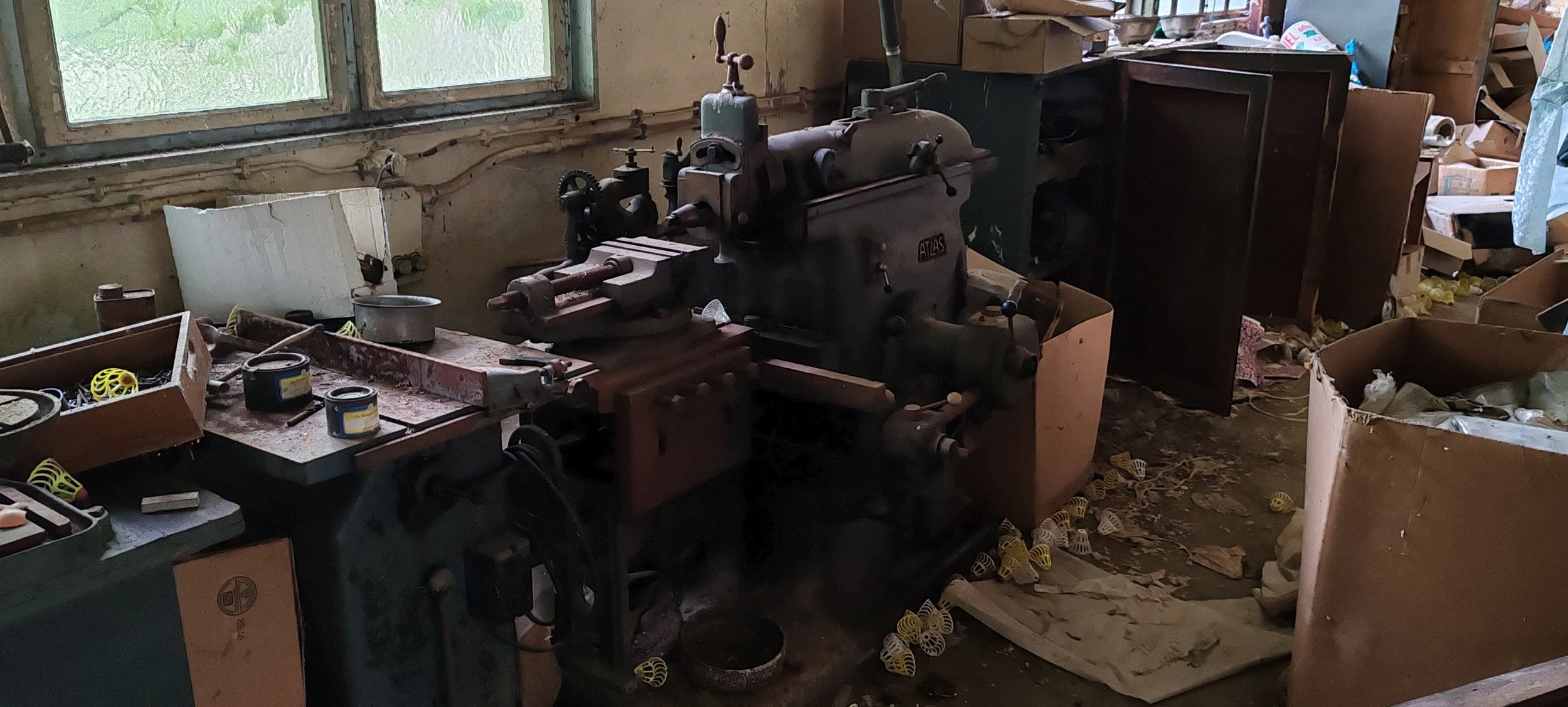 Ein alter Betrieb in einem vernachlässigten Raum mit Maschinen, Staub und herumliegenden Gegenständen, beschädigter Fensterfront.