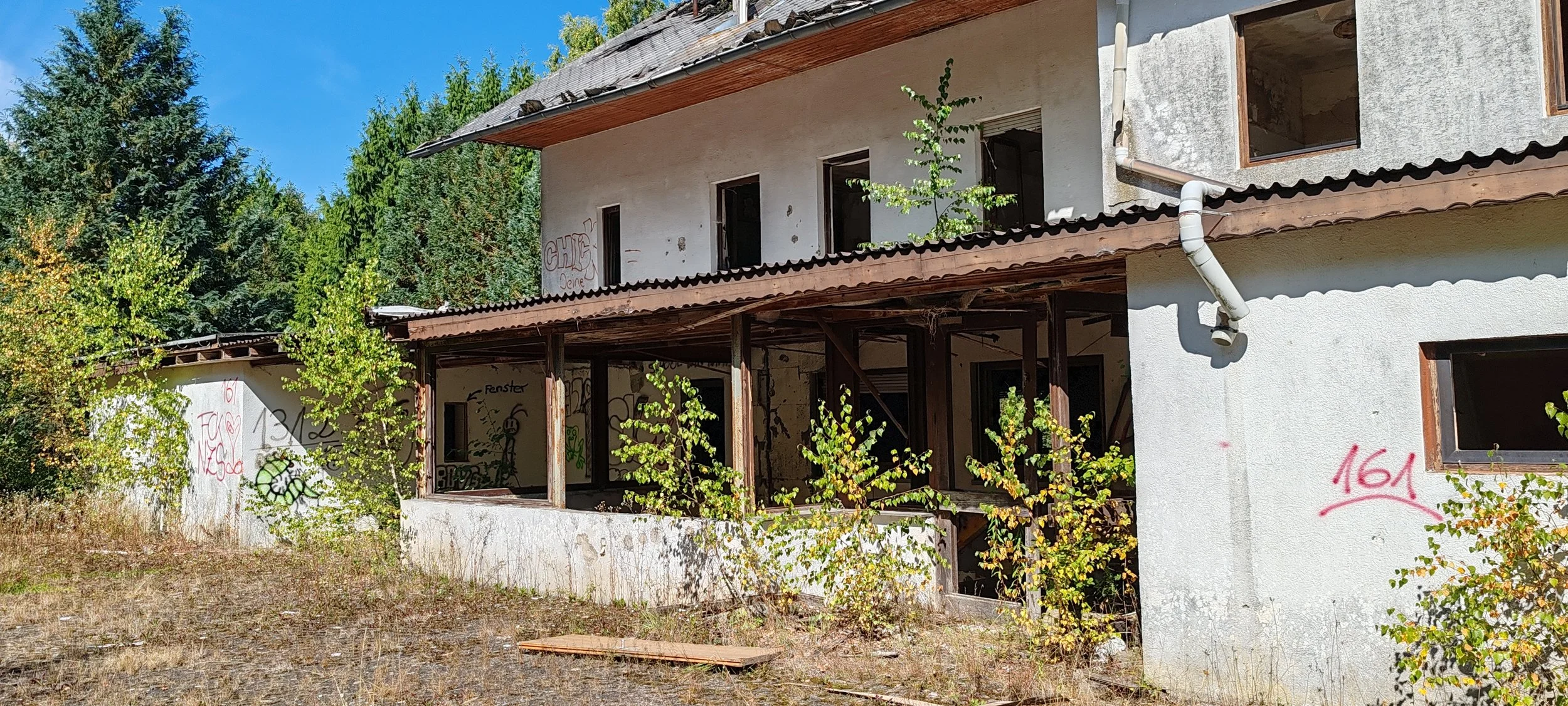 Verfallenes, zweistöckiges Gebäude mit Graffiti an den Wänden, bröckelnde Putzschichten, offene Fenster und Überdachung, umgeben von Büschen und Bäumen, auf einer trockenen, grasigen Fläche unter blauem Himmel.