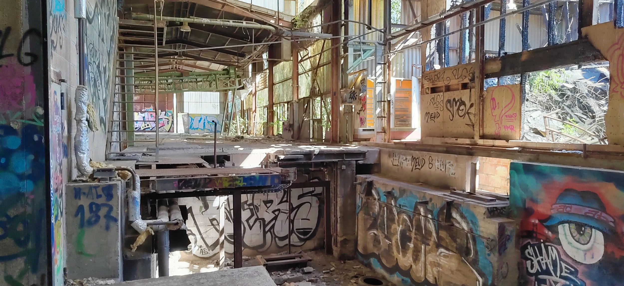 Ein verlassener, mit Graffiti bemalter Gebäudeinnerraum mit Holz- und Metallstrukturen, teilweise zerfallenen Böden und offenen Fenstern, durch die natürliches Licht einfällt.