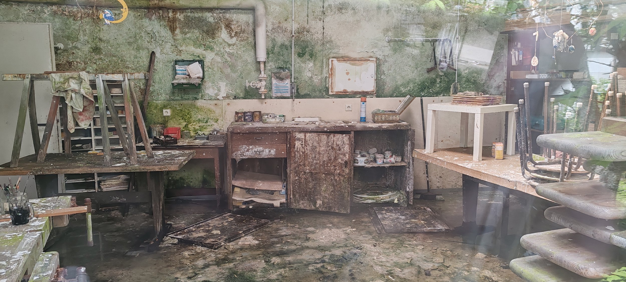 Ein vernachlässigtes, schmutziges und verrostetes Atelier mit beschädigten Möbeln, schmutzigem Boden und moosbedeckten Wänden.