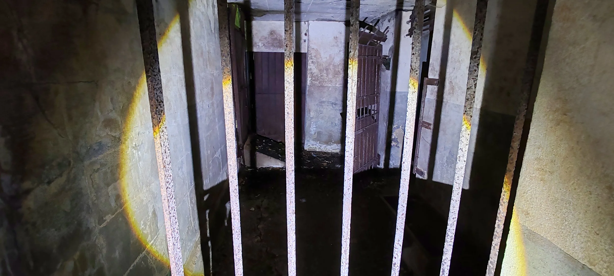 Eine verlassene, dunkle Zelle mit rostigen Gitterstäben und schmutzigem Boden innerhalb einer alten Steinmauer.