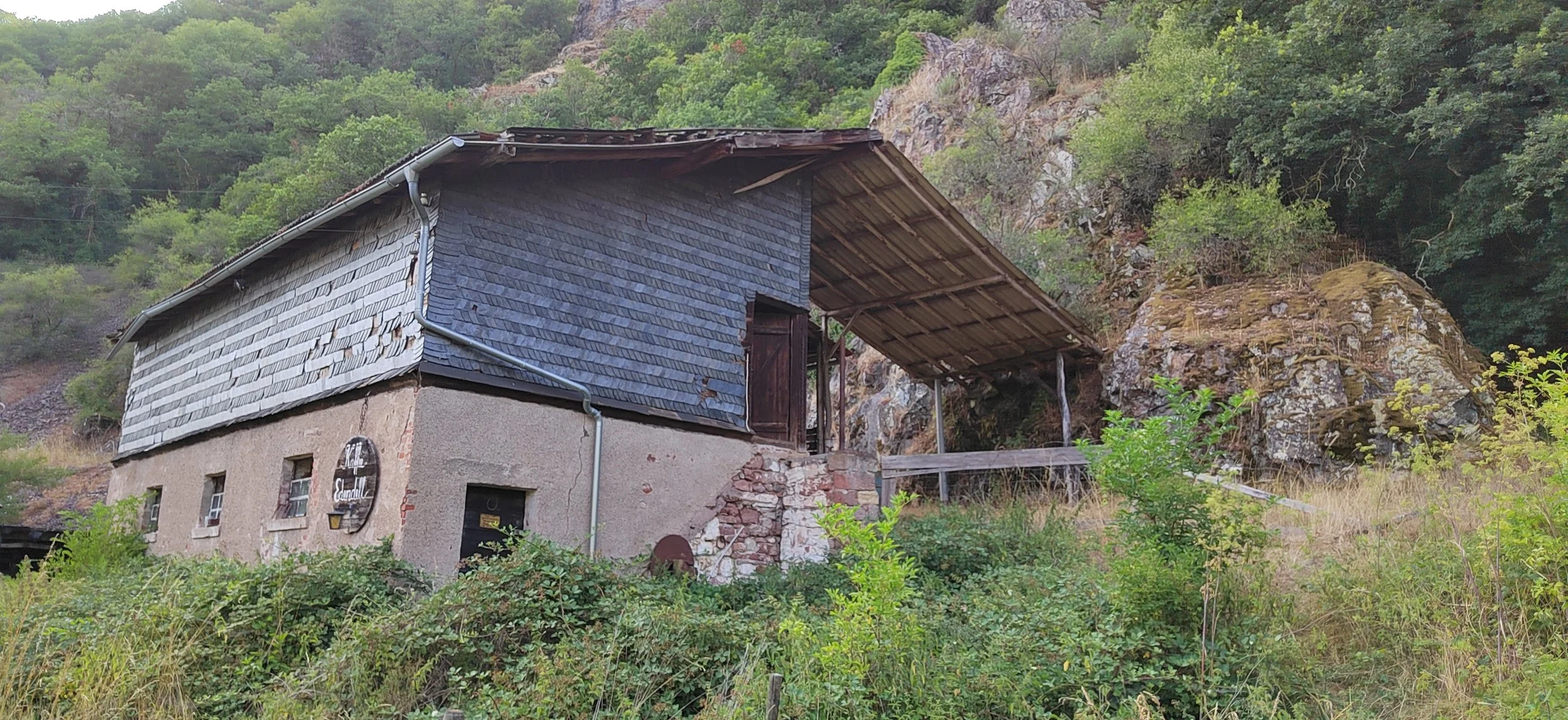 Altes Haus auf einer Hügel inmitten von Büschen und Bäumen, mit Steinen im Hintergrund.