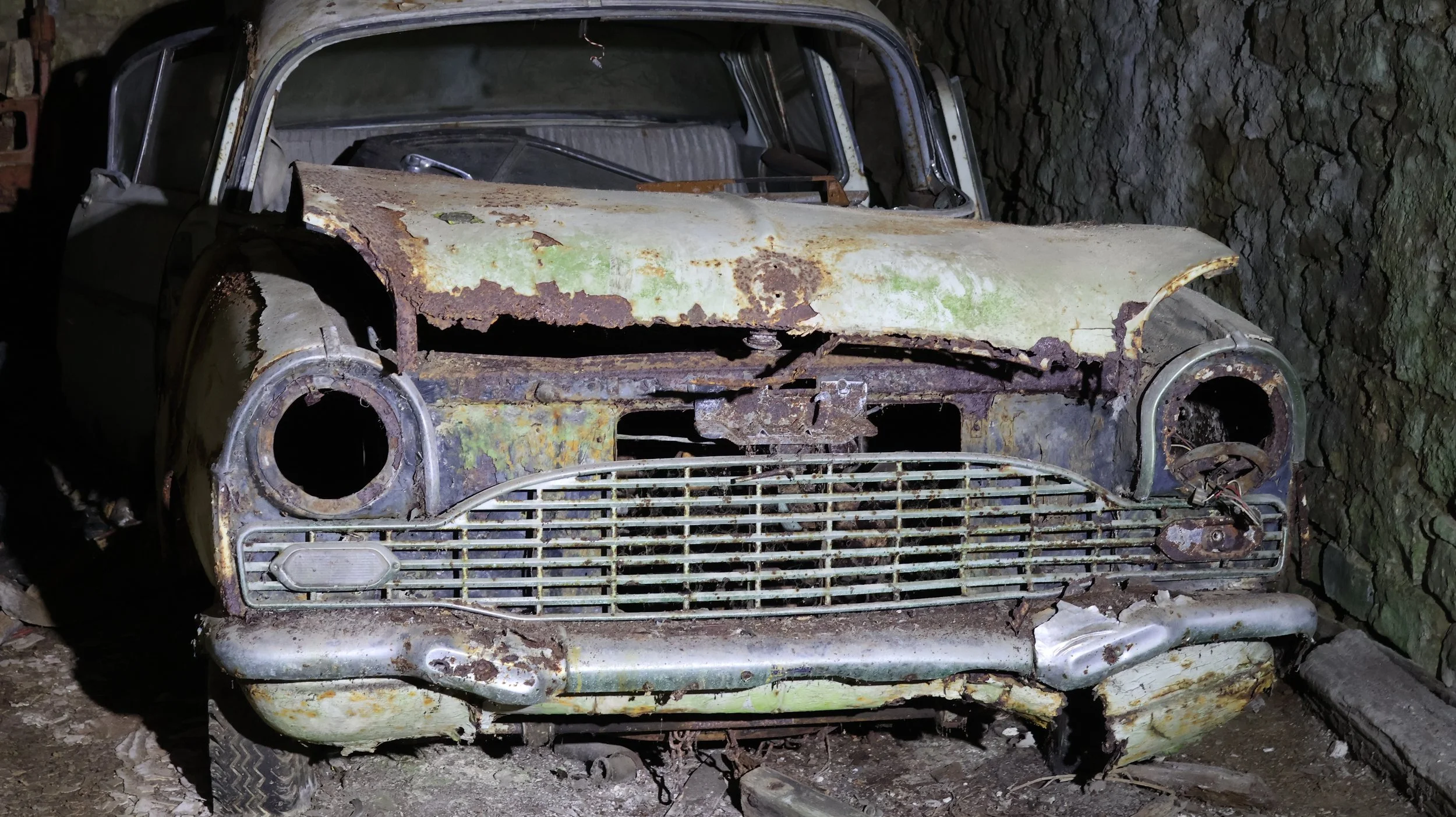 Ein verlassenes, verrostetes Auto mit zerbrochener Frontscheibe, das in einer dunklen Höhle oder Garage steht.