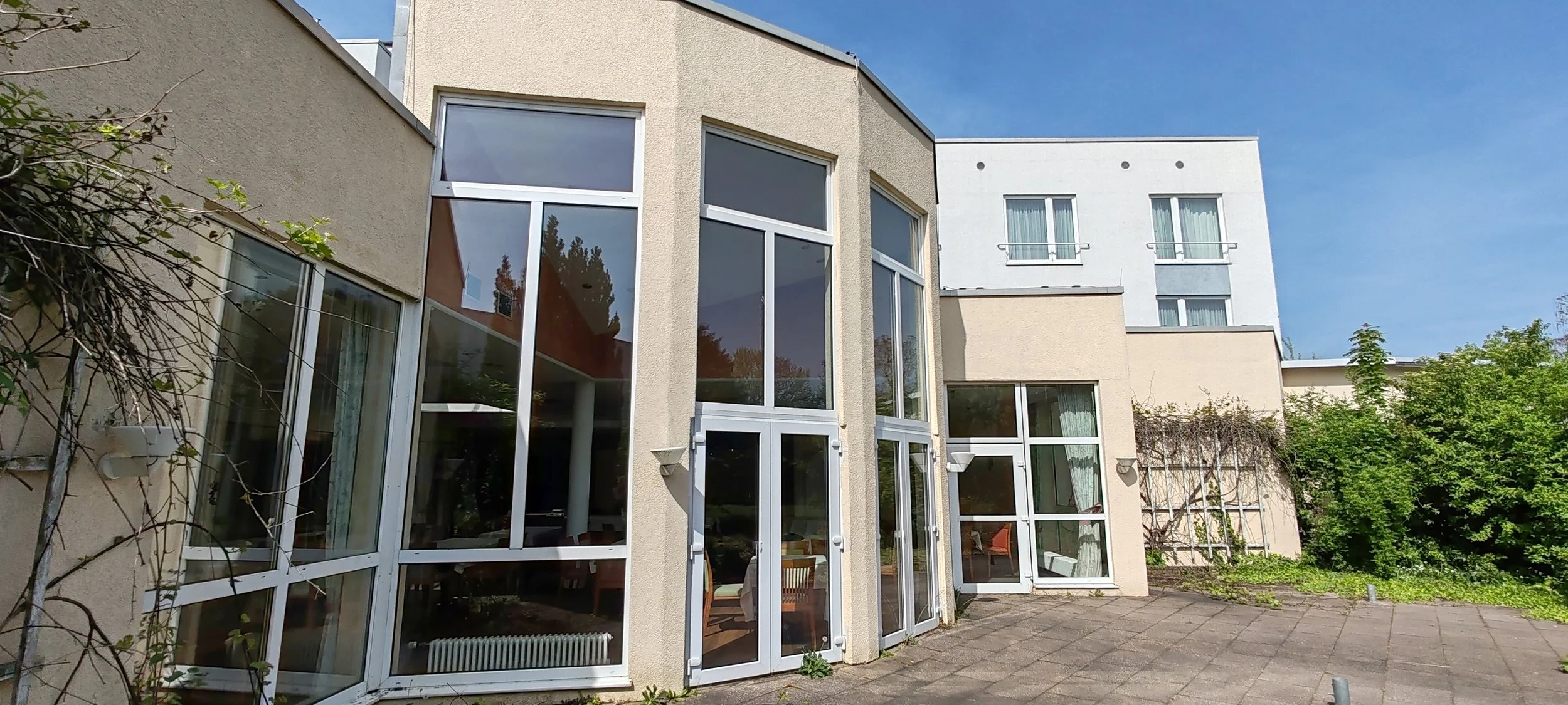 Mehrstöckiges Gebäude mit großen Fenstern und Terrasse, umgeben von Pflanzen, bei sonnigem Himmel.