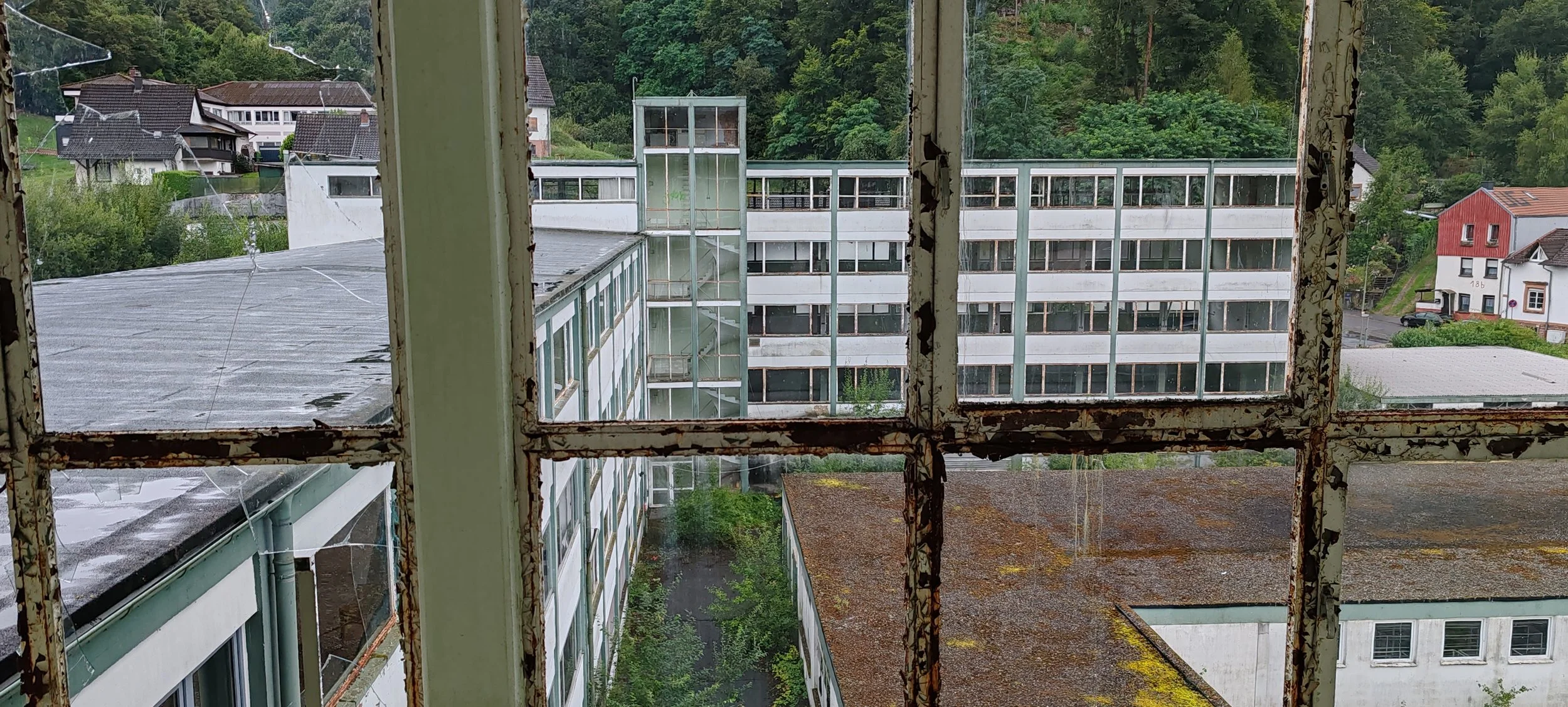 Blick aus einem alten, verrosteten Fenster auf ein modernes, mehrstöckiges Gebäude mit vielen Fenstern, umgeben von grüner Natur und einigen Häusern.