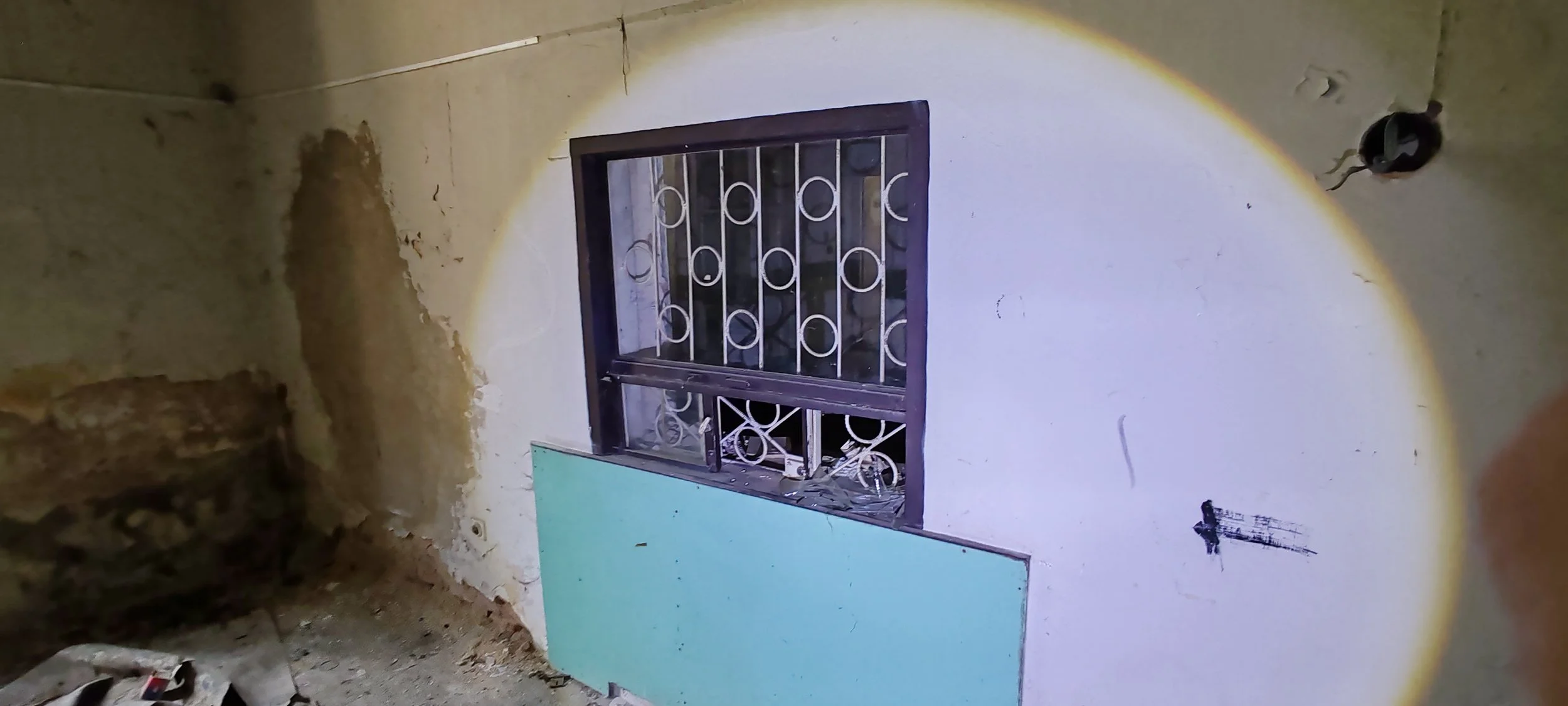 Ein Fenster mit Metallgitter an einer alten Wand, die teilweise beschädigt ist, im Inneren eines Raumes, der offenbar renoviert wird.