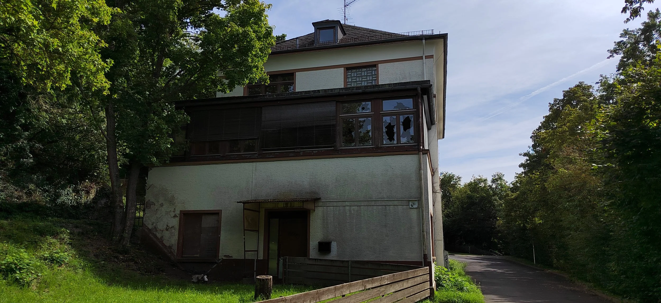 Mehrstöckiges Haus mit dunklen Fensterläden, umgeben von Bäumen, in einer ruhigen, bewaldeten Gegend, bei Tageslicht.