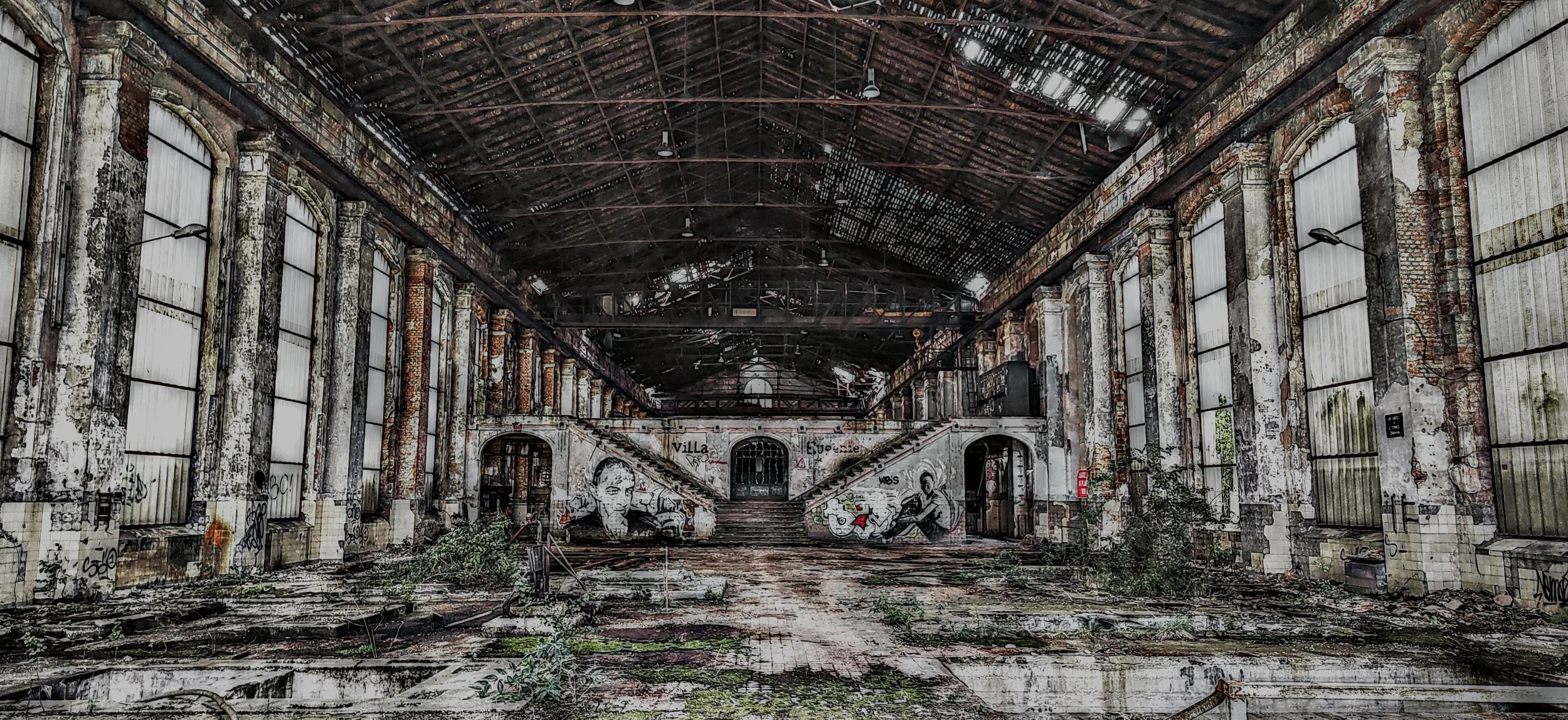 Verlassene, verfallene Industriehalle mit Graffiti an den Wänden, zerfallendem Dach und überwachsener Bodenfläche.