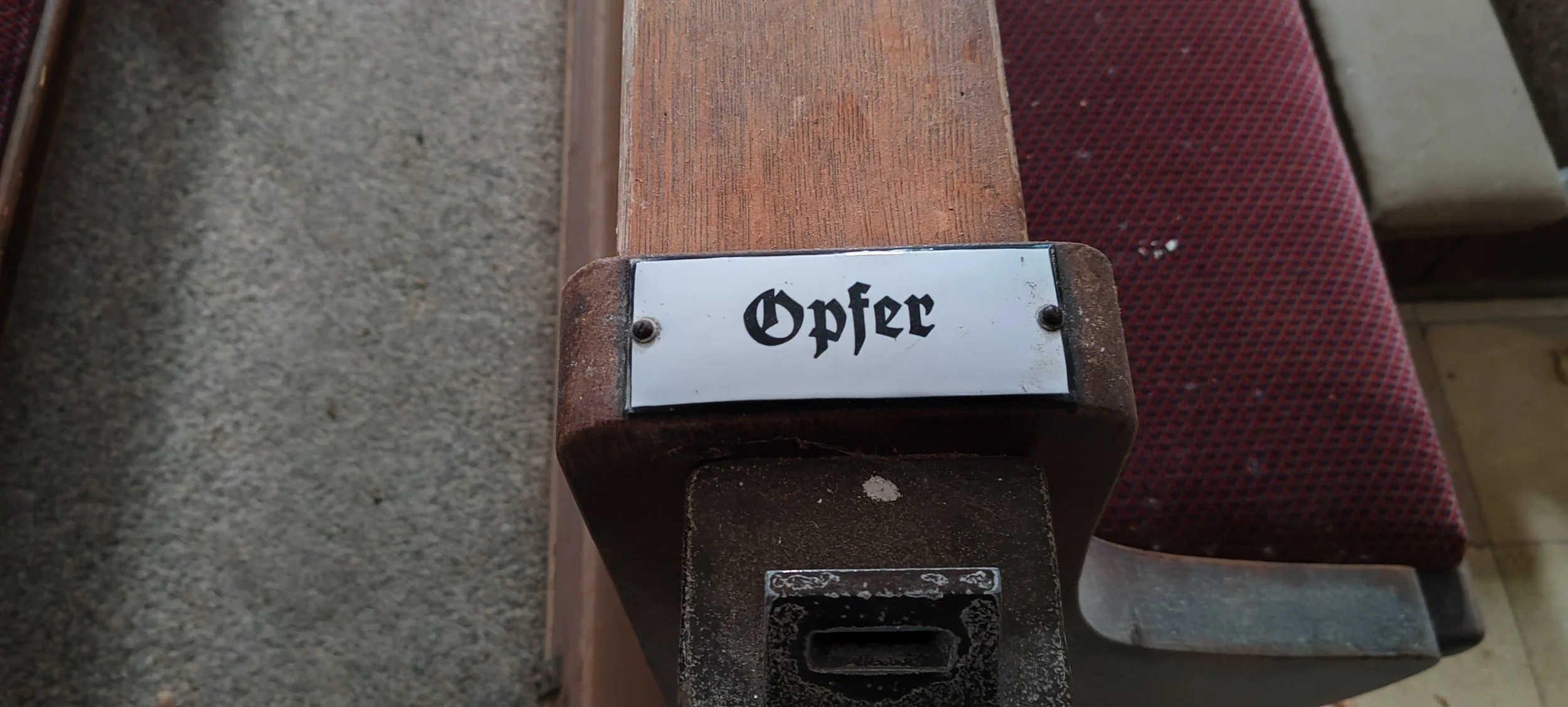 Ein altes, rustikales Holzstück mit einer Metallplakette, auf der das Wort 'Opfer' in gotischer Schrift steht, und die auf einer dunklen, wahrscheinlich alten Maschine oder einem Gegenstand befestigt ist.