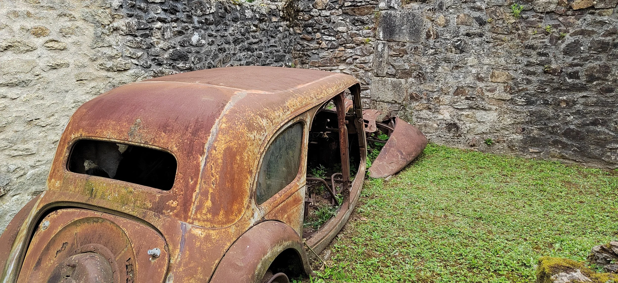 Verrostetes, altes Auto auf einer Wiese neben einer Steinmauer, umgeben von Grün und Moos.