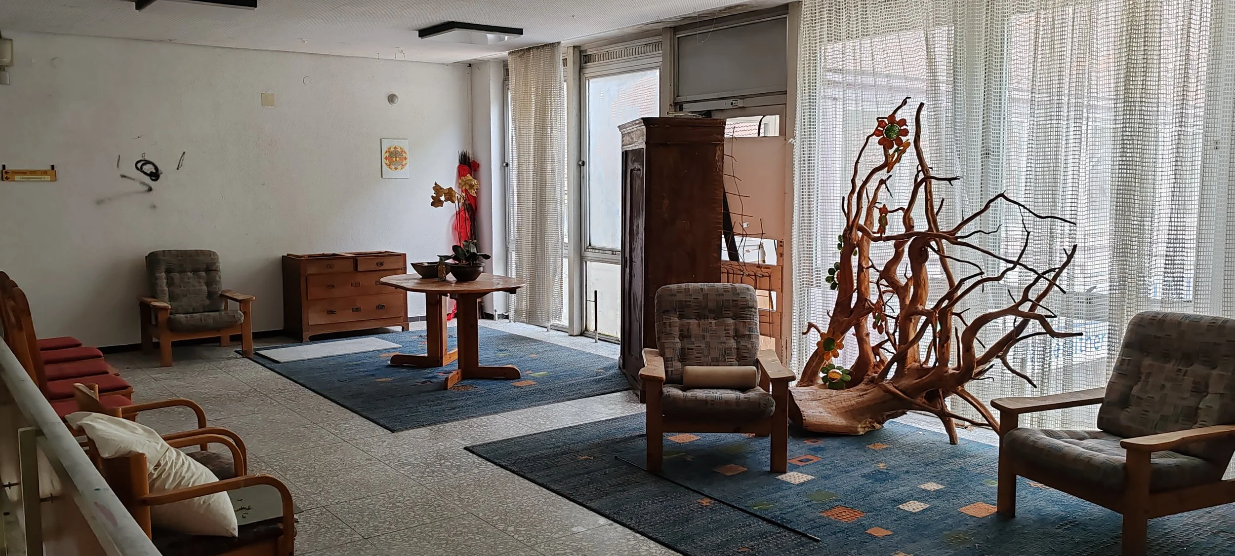 Ein Aufenthaltsraum mit mehreren Sesseln und Tischen, einer großen Holzskulptur eines Baumes an der Fensterseite und Vorhängen vor den Fenstern, die Tageslicht hereinlassen.