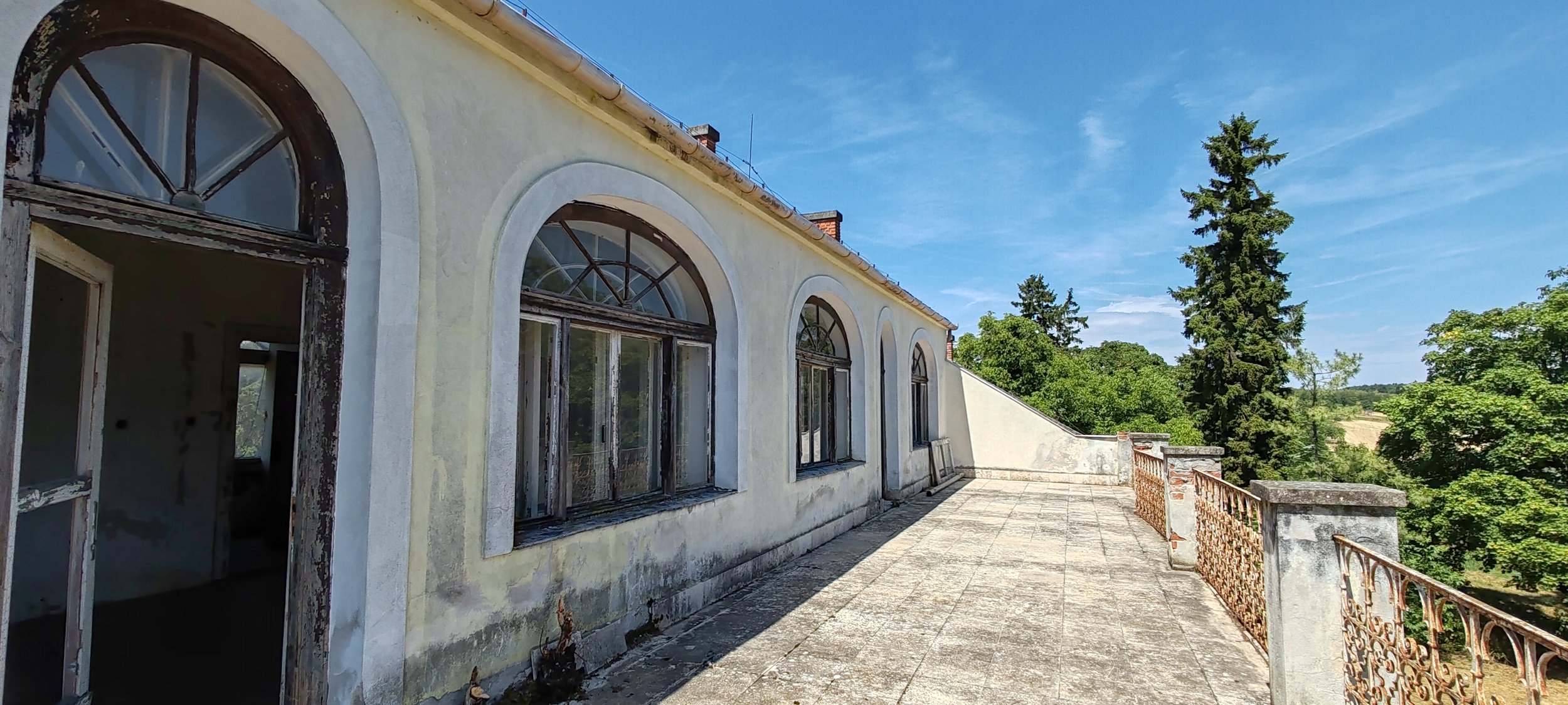 Verlassene Terrasse an einem alten Haus mit großen Fenstern und einer Eisenbrüstung, umgeben von Bäumen bei sonnigem Himmel.
