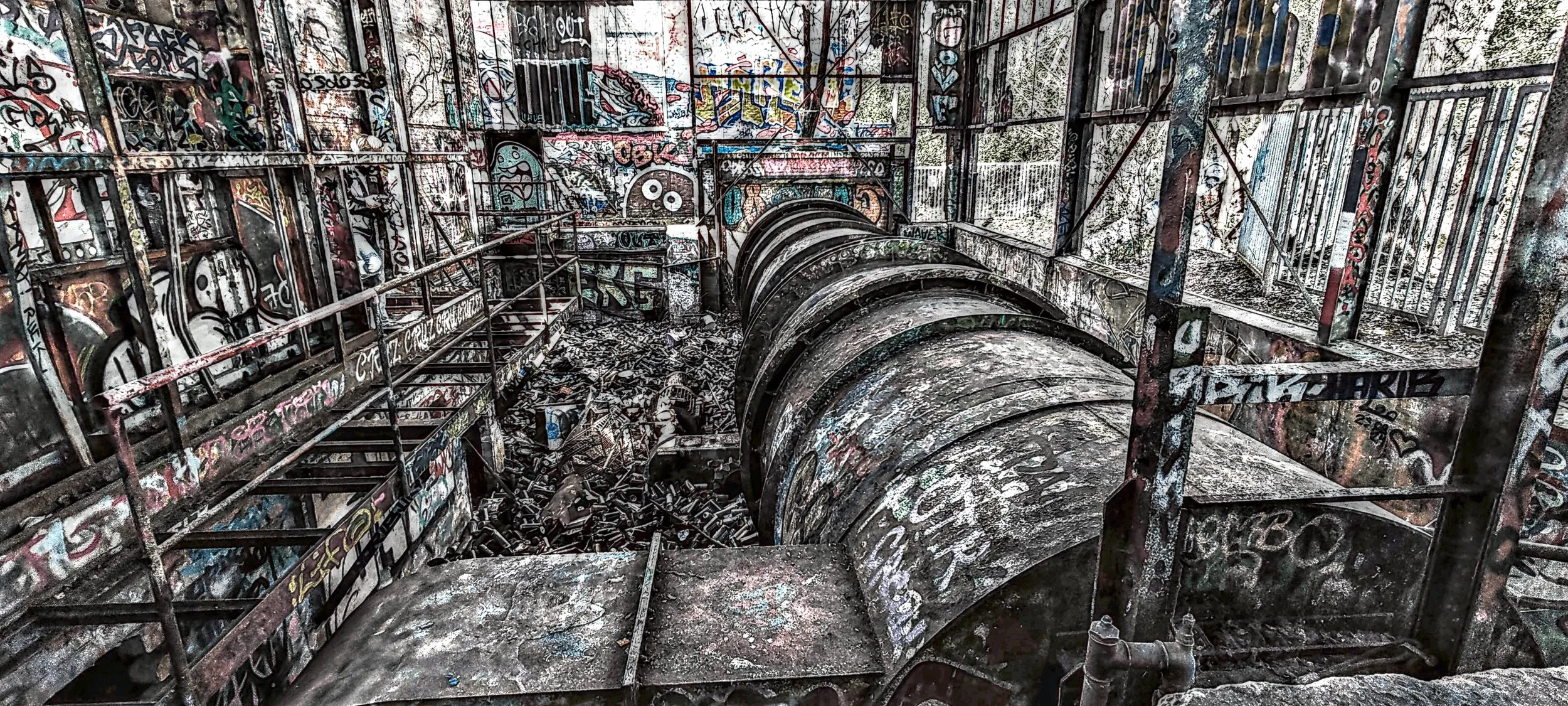 Ein verfallener, mit Graffiti bedeckter, industrieller Ort mit einem großen, rostigen, Spiralrohr und einer umlaufenden Metallsicherung. Überall liegen Trümmer und Schrott, und das Umfeld wirkt verlassen und verwahrlost.