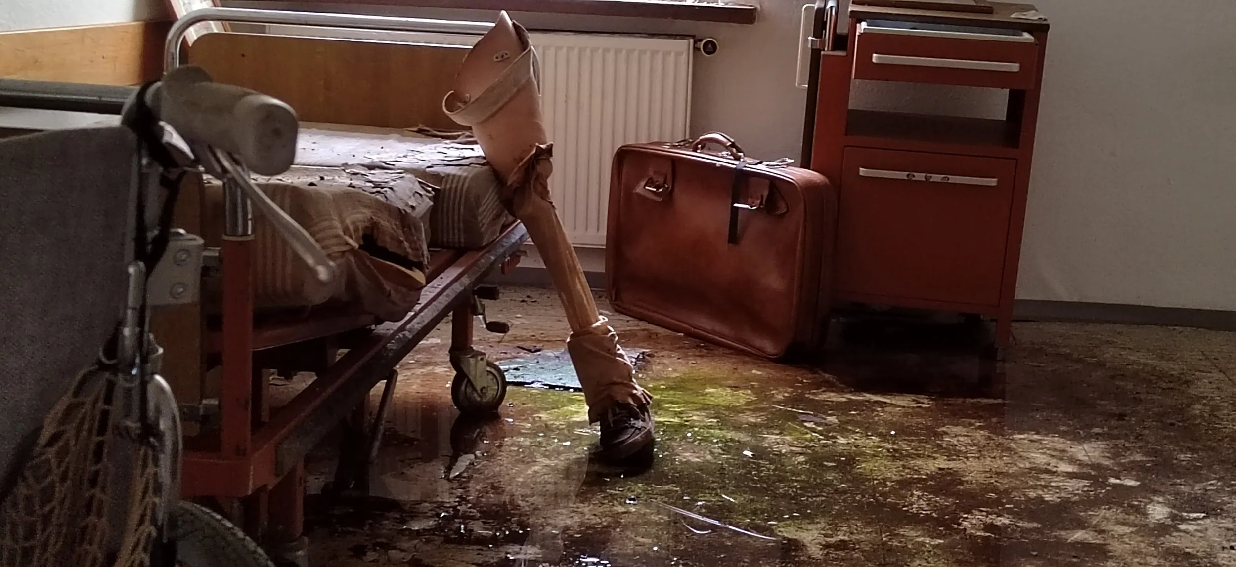 Ein veraltetes Krankenzimmer mit einem vollgelaufenen Boden, einem kaputten Bett, einem großen braunen Koffer, einem kleinen -schrank und einer Radrennmaschine.