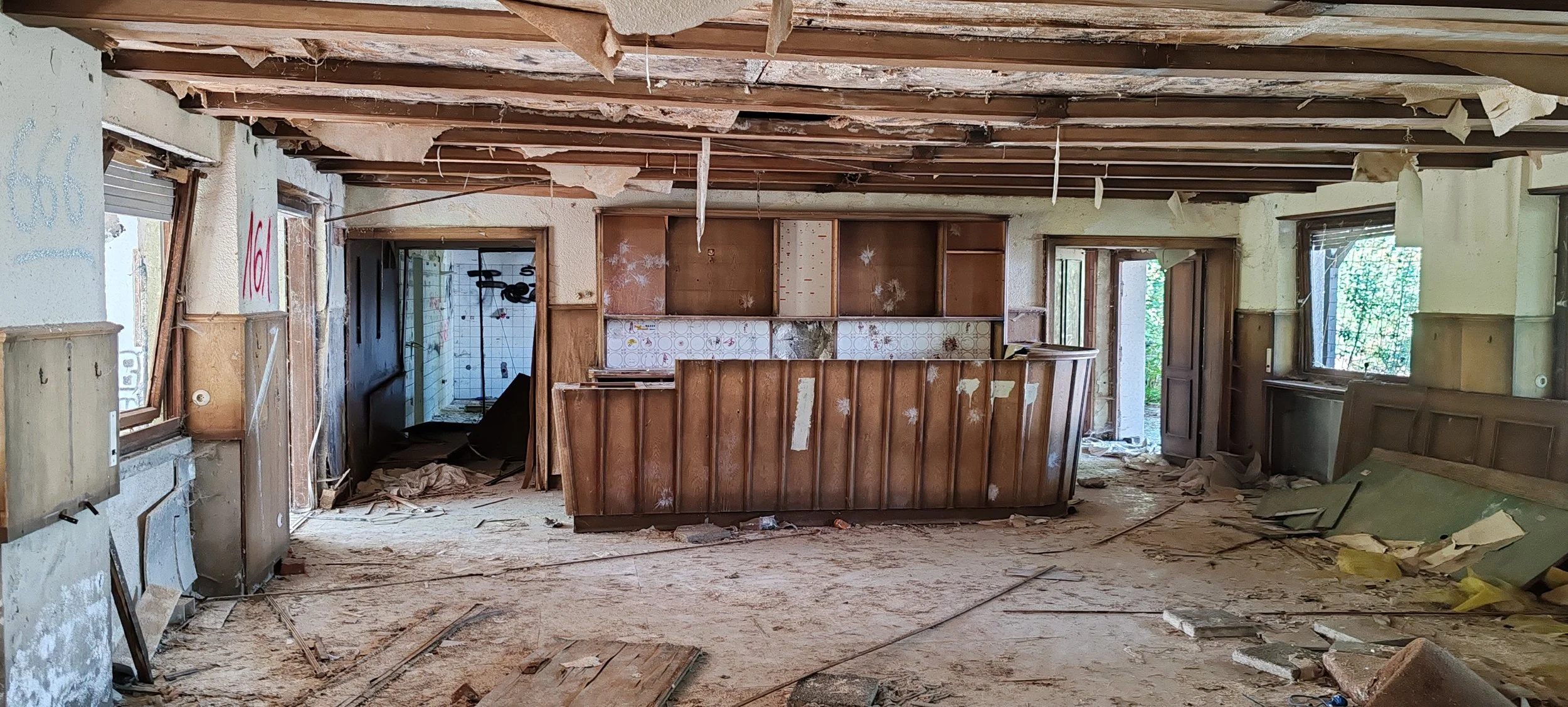 Innenansicht eines verlassenen, baufälligen Hauses mit offenem Deckenbalken und gegenüberliegenden Fenstern und Türen, umgeben von Schmutz, Staub und zerbrochenem Material.