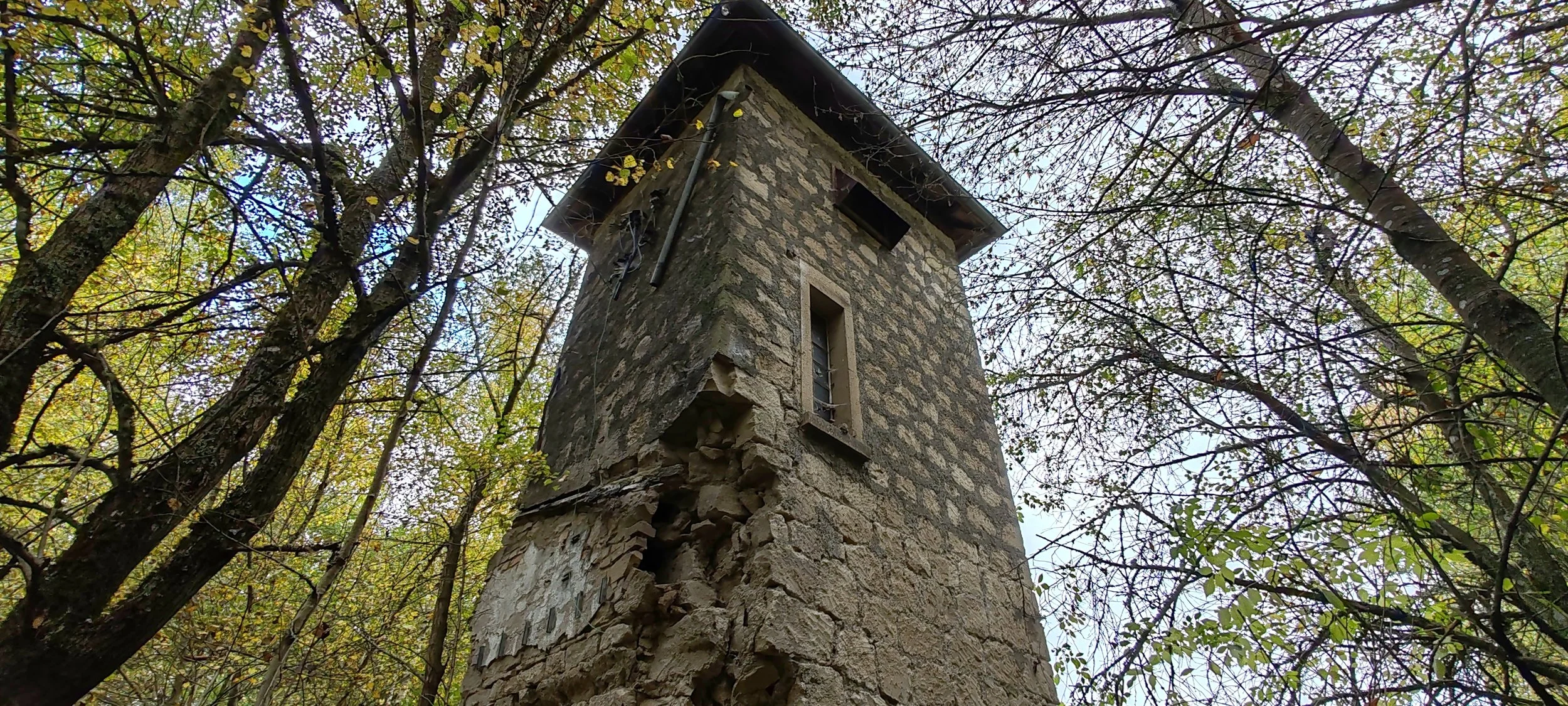 alter, zerfallener Turm aus Steinen in einem Wald, mit kleinen Fenstern und einem schrägen Dach, umgeben von Bäumen mit Herbstlaub.