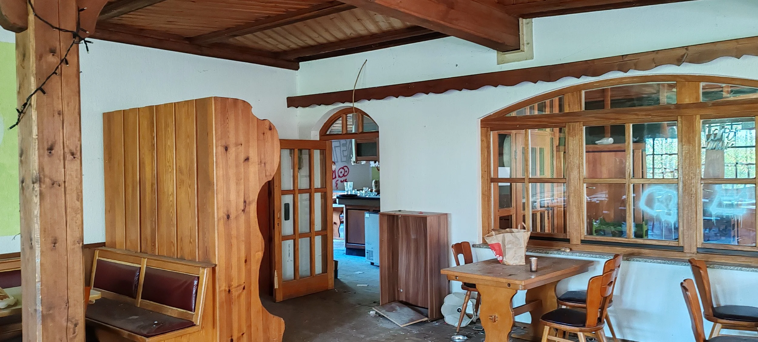 Innenraum eines rustikalen Hauses mit Holzinterieur, Ess- und Wohnzimmerbereich, Holzstühlen, einem Tisch mit Papiertüte und Kerze, großen Fenster mit Holzrahmen.