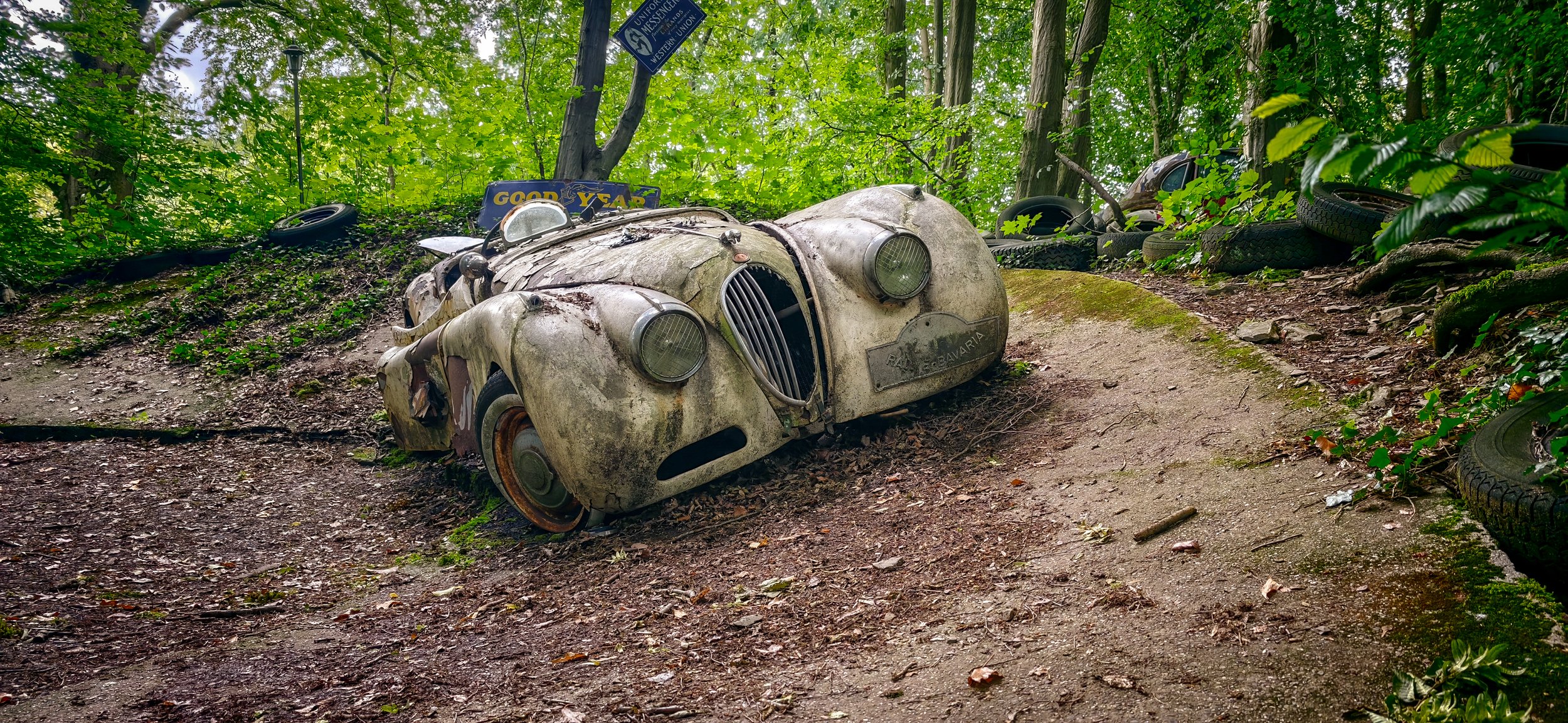 Ein verrosteter, verfallener alter Rennwagen in einem Wald, umgeben von Bäumen, Reifen und einem Goodyear-Schild.
