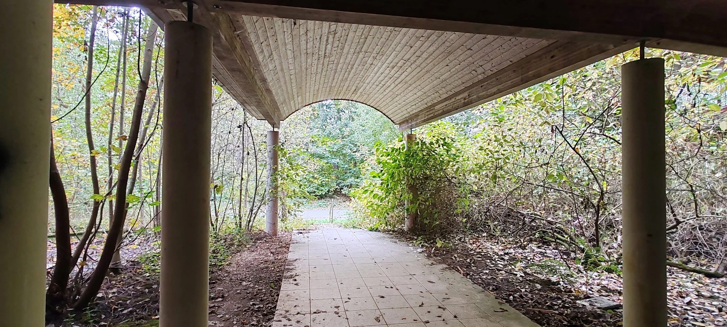 Unterführungsbogen mit Säulen, der Weg führt ins Grüne, umgeben von Büschen und Bäumen.