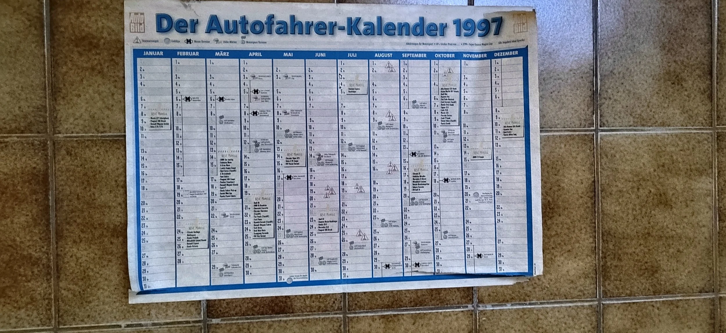 Ein Wandkalender für Autofahrer aus dem Jahr 1997, monatsweise aufgeteilt, mit Markierungen und Notizen, an einer Fliesenwand befestigt.