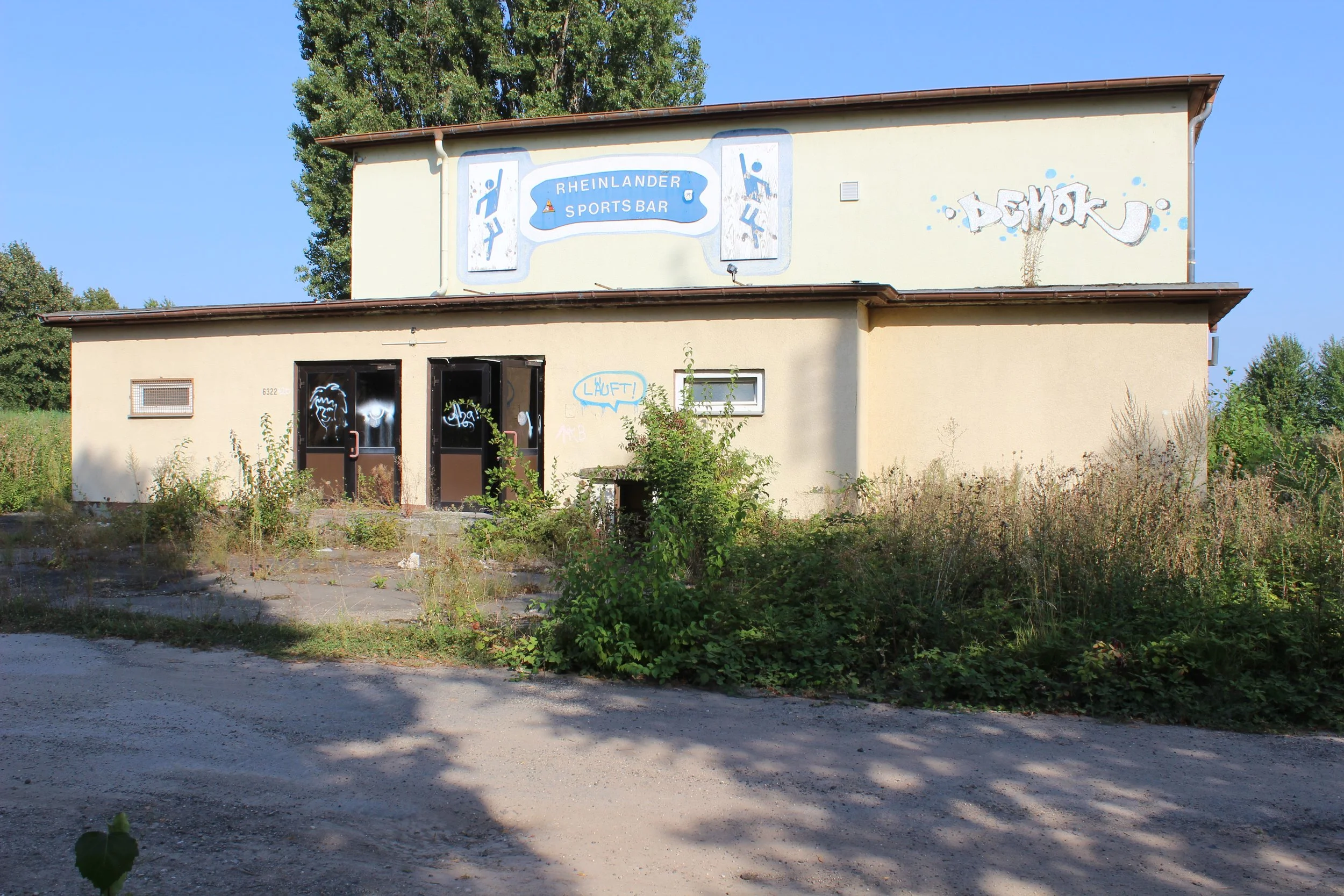 Verfallenes Gebäude mit Graffiti, das den Rheinländer Sports Bar bewirbt, umgeben von wildem Gras und Büschen, im Freien, bei Sonnenschein.