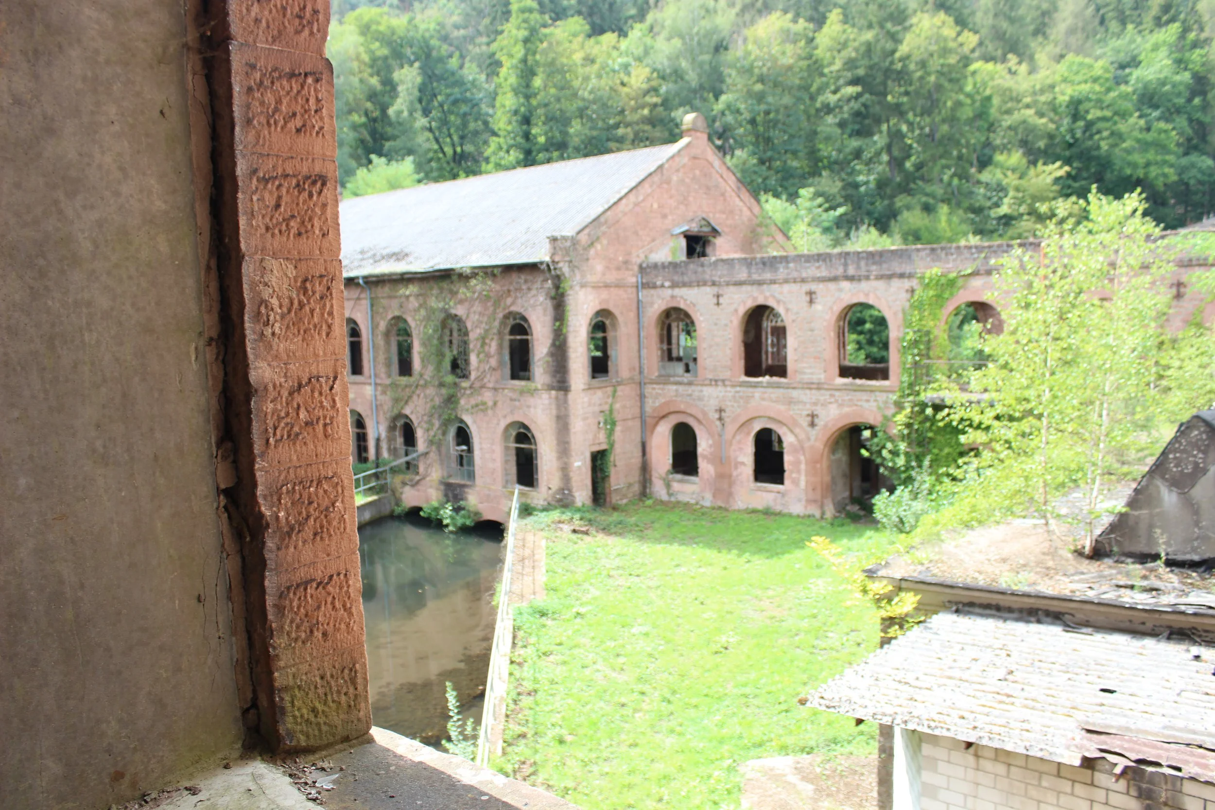 Blick auf eine alte, verlassene Kirche mit Bogenfenstern, umgeben von Bäumen und grünem Garten, gesehen aus einem offenen Fenster.