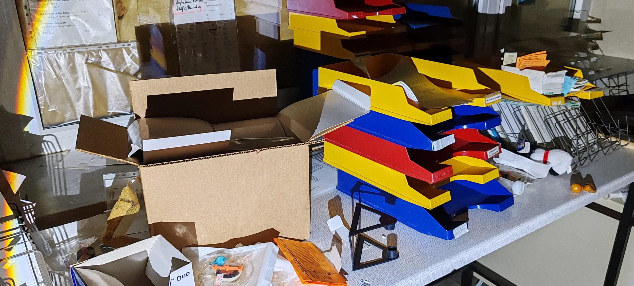 Bürobedarf, insbesondere Farbkarten in den Farben Rot, Blau, Gelb und Schwarz, auf einem Tisch. Es gibt auch leere und gefüllte Verpackungsschachteln, Papier, einen Einkaufswagen, und kleine Gegenstände.