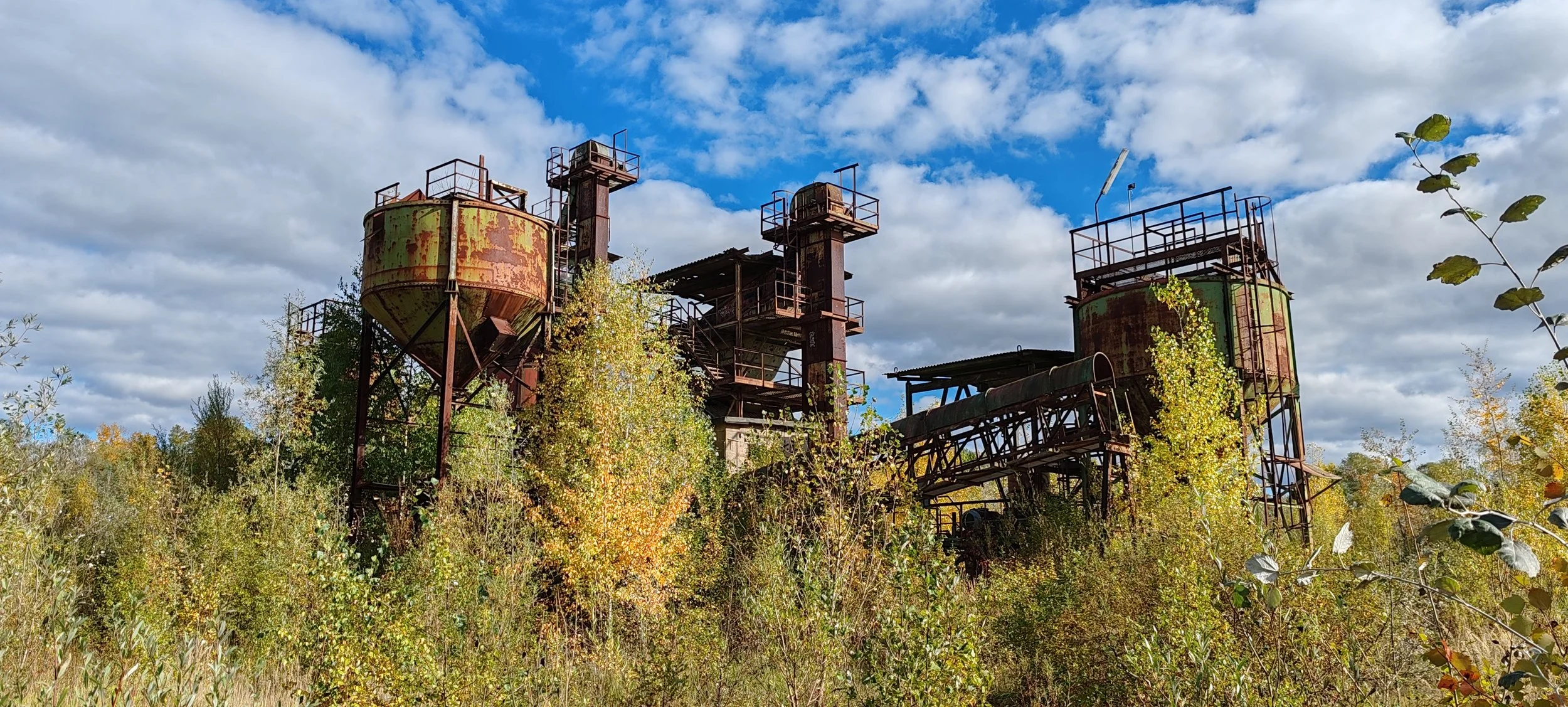 Verfallene, rostige Industrieanlagen im Grünen unter einem bewölkten Himmel.