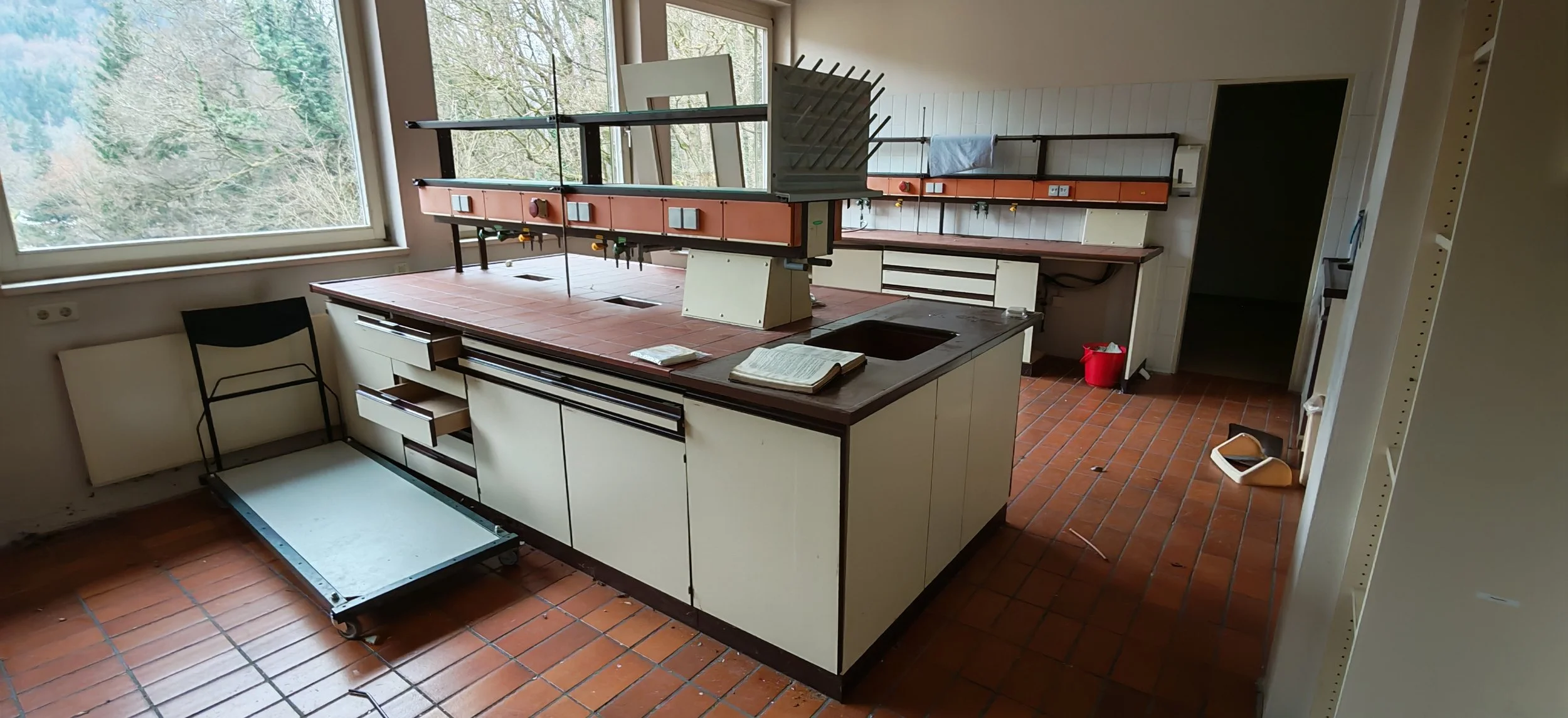 Leeres, veraltetes Küchenlabor mit zentralem Arbeitstisch, großen Fenstern, offenen Schubladen, einem Buch und einem roten Müllkorb auf dem Boden.