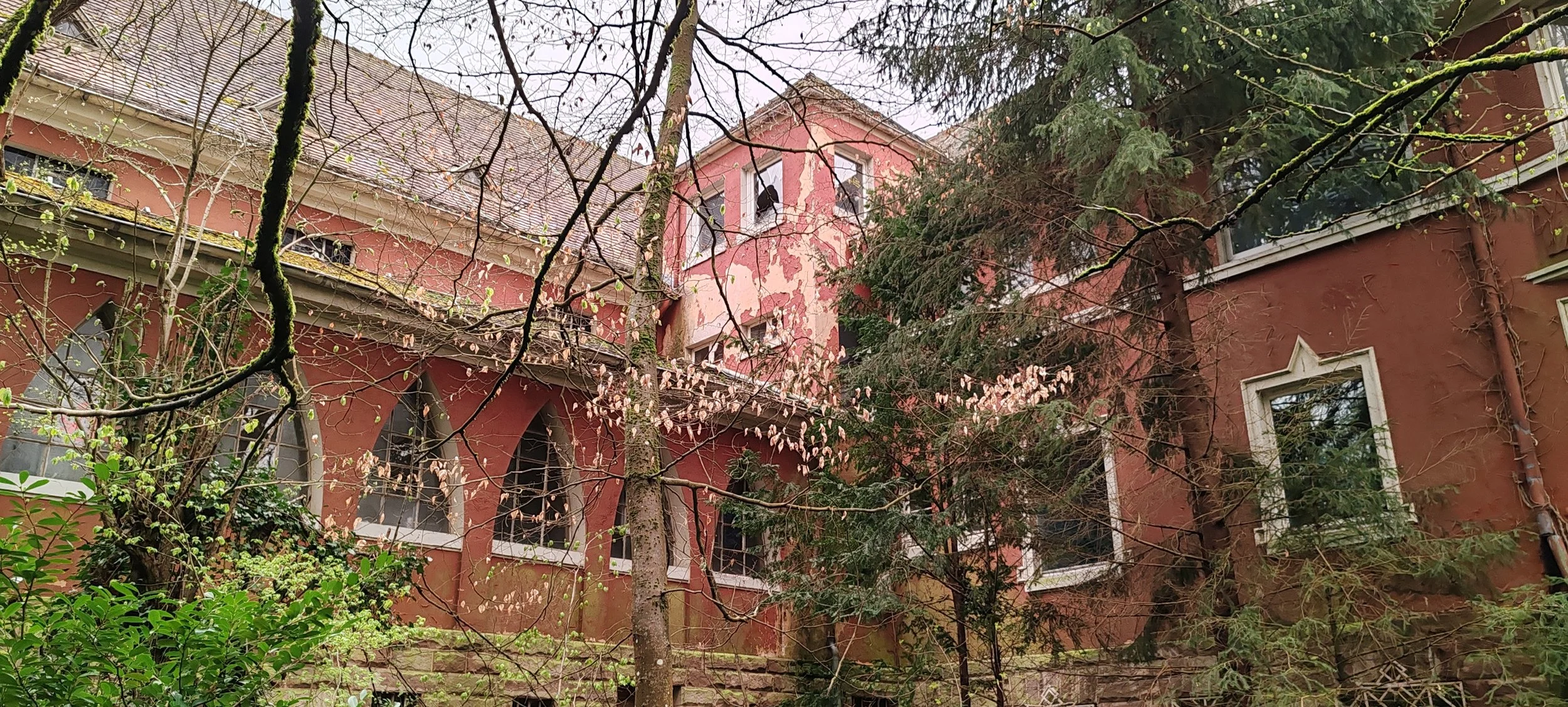 Ein altes, rotes Gebäude mit mehreren Fenstern, umgeben von Bäumen und Sträuchern im Frühling.