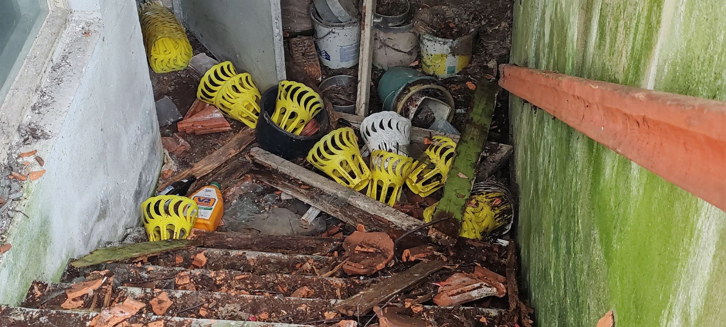 Baustellenabfälle und Müll, darunter gelbe und weiße Plastikkegel, Schutt, Ziegelreste, Werkzeugteile, und eine Flasche mit Sandpapier in einem engen Raum zwischen grüner und weißer Wand.