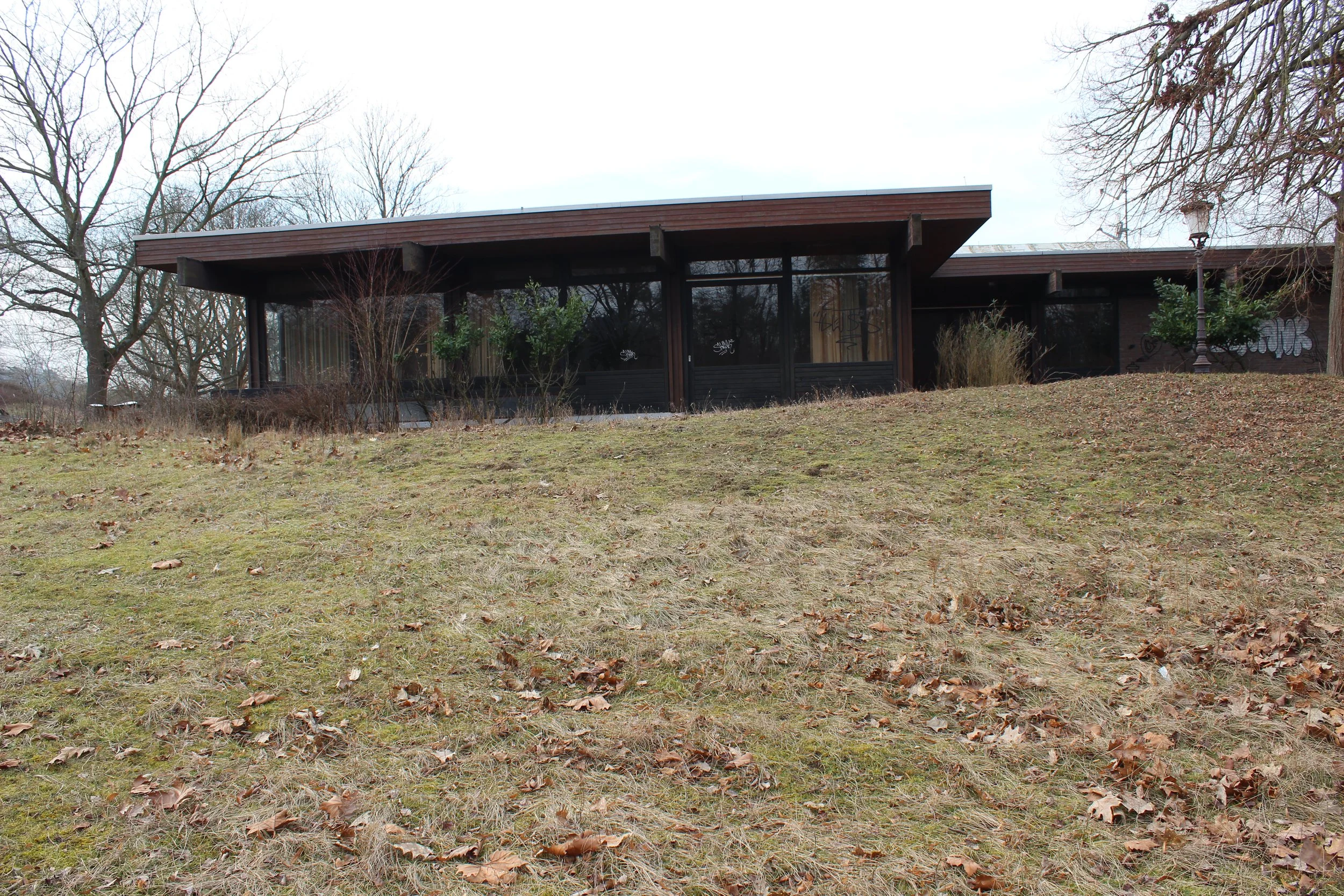 Ein modernes, flaches Gebäude mit großen Fenstern, umgeben von kahlen Bäumen und flachem Gras, das im Frühling oder Herbst liegt, bei bewölktem Himmel.