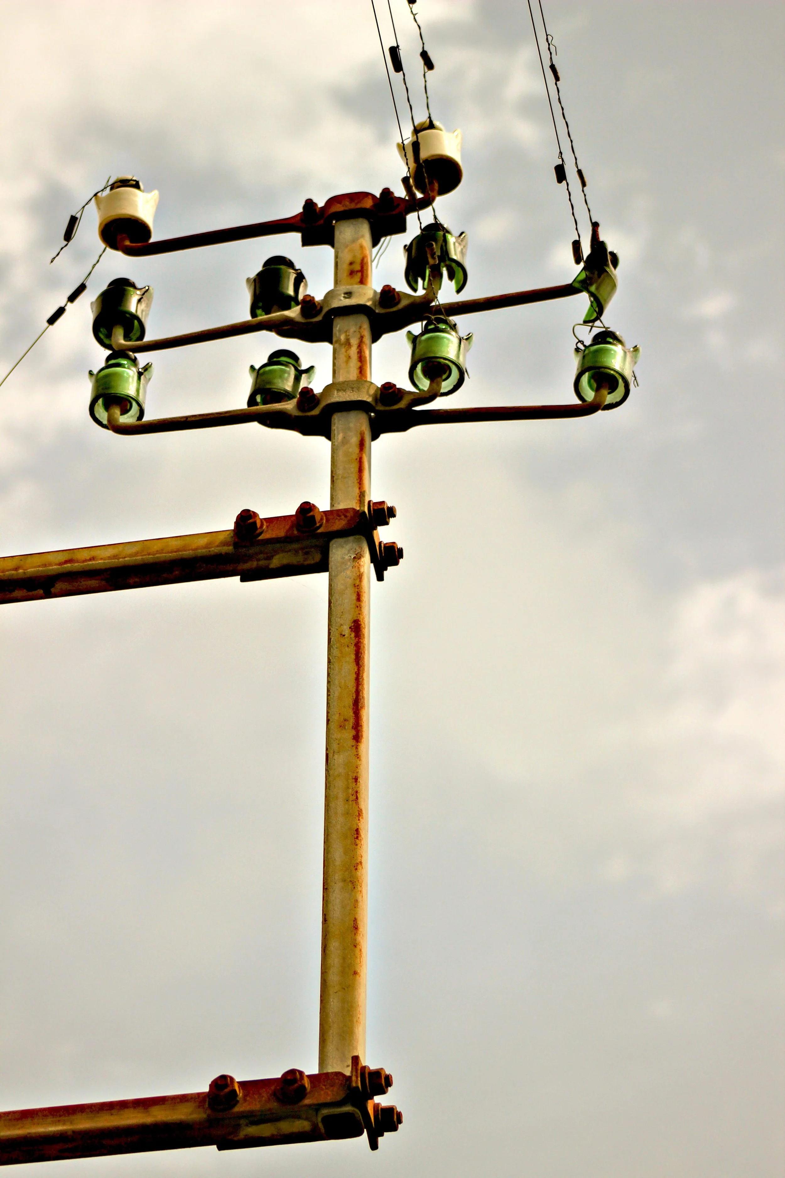 Ein verrosteter Strommast mit mehreren grünen Isolatoren unter einem bewölkten Himmel.