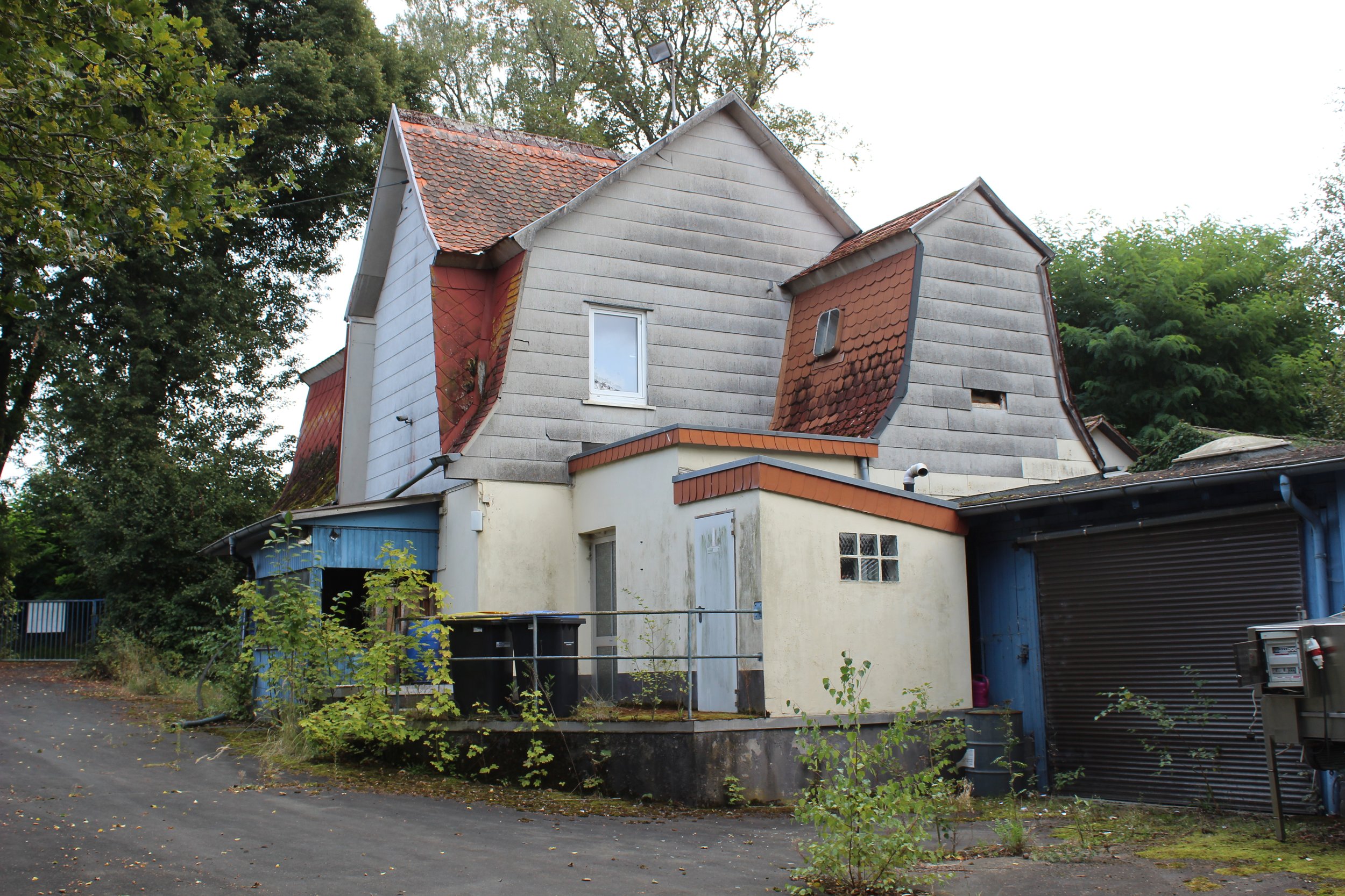 Ein altes, abgängiges Haus mit ungleichmäßigen Dächern, teilweise mit roten Ziegeln gedeckt, und einer kleinen, modernen Erweiterung im Vordergrund. Es steht neben einer Straße, umgeben von Bäumen und Bewuchs, mit Rolltoren und Mülltonnen im Vordergr