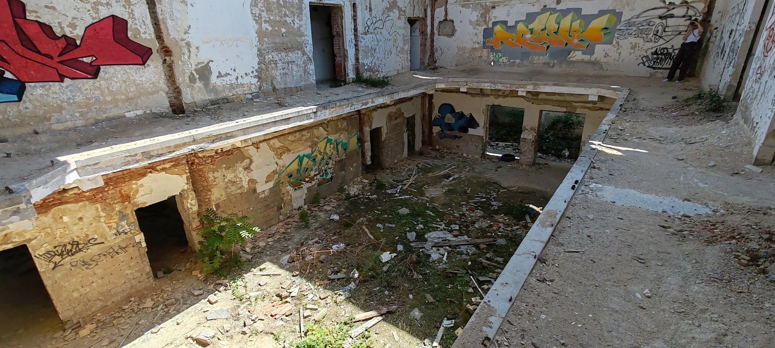 Verlassener, ruinöser Raum mit Graffiti an den Wänden, Boden voller Schutt und Pflanzen, und eine Rundung, die auf einen ehemaligen Swimmingpool oder Wasserbecken hindeutet.