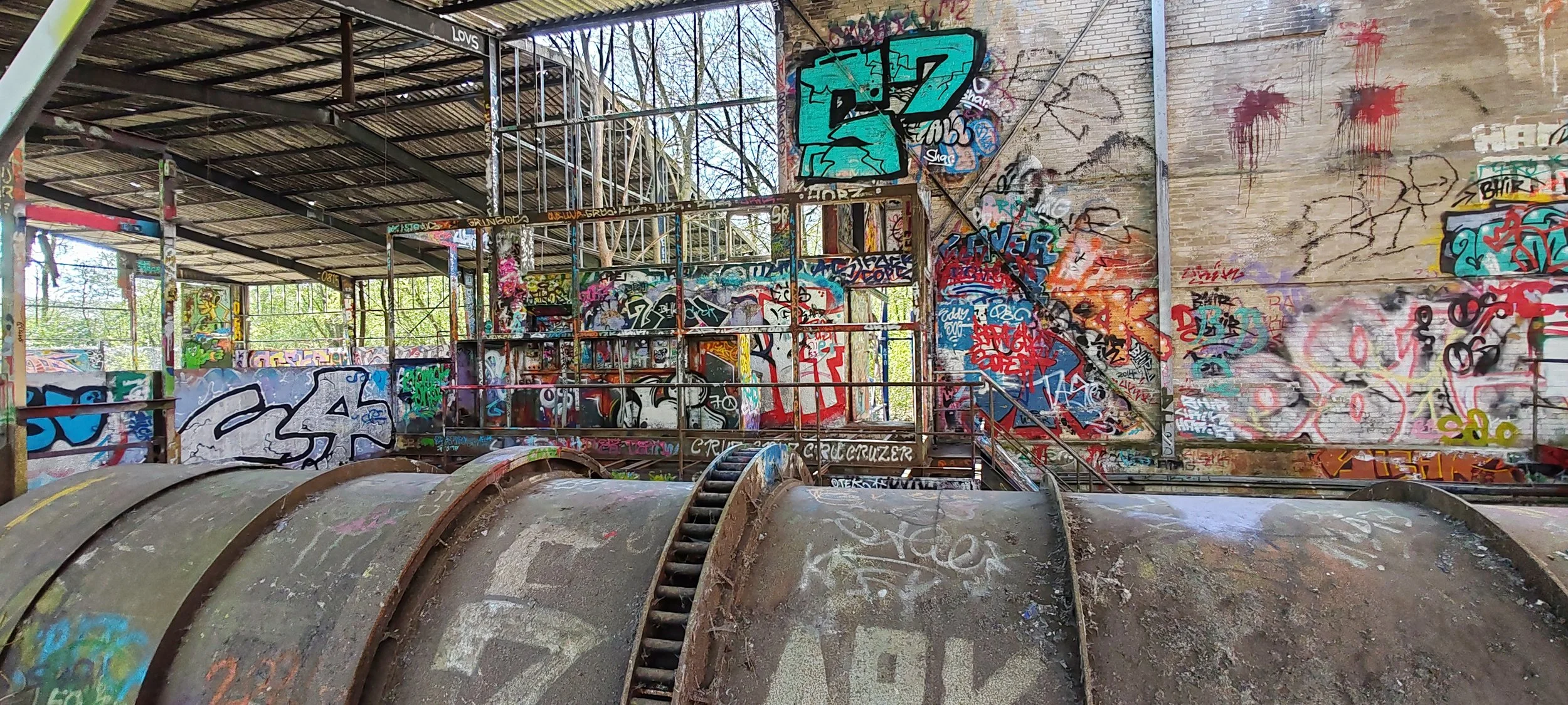 Verfallenes Gebäude mit Graffiti bedeckten Wänden, Ruinen, Stahltreppen und einer großen, rostigen runden Struktur im Vordergrund.
