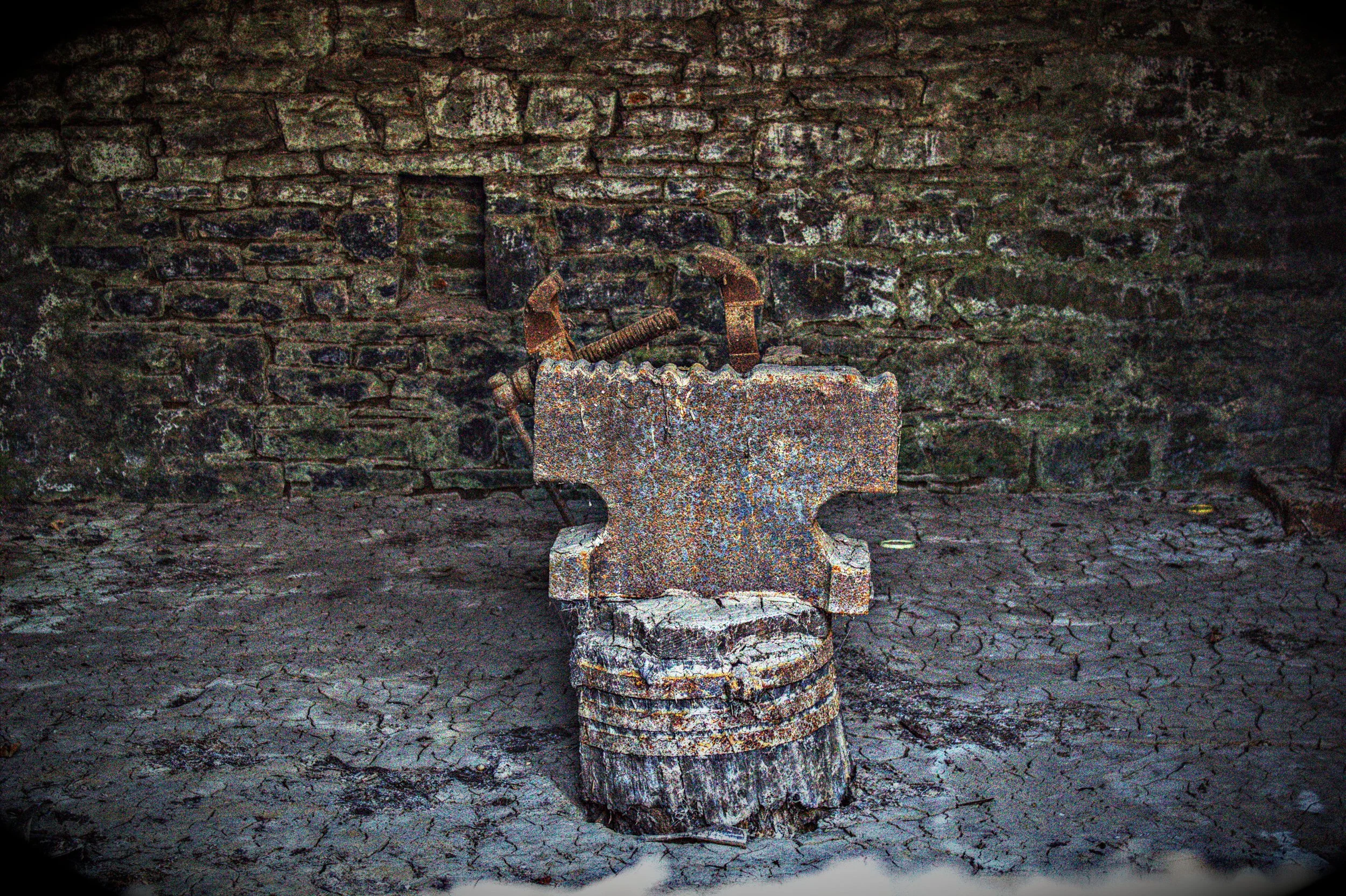 Alte, verrostete Amboss mit Hammer in einer alten Werkstatt oder Schmiede, umgeben von einer Steinmauer und einem trockenen, rissigen Boden.