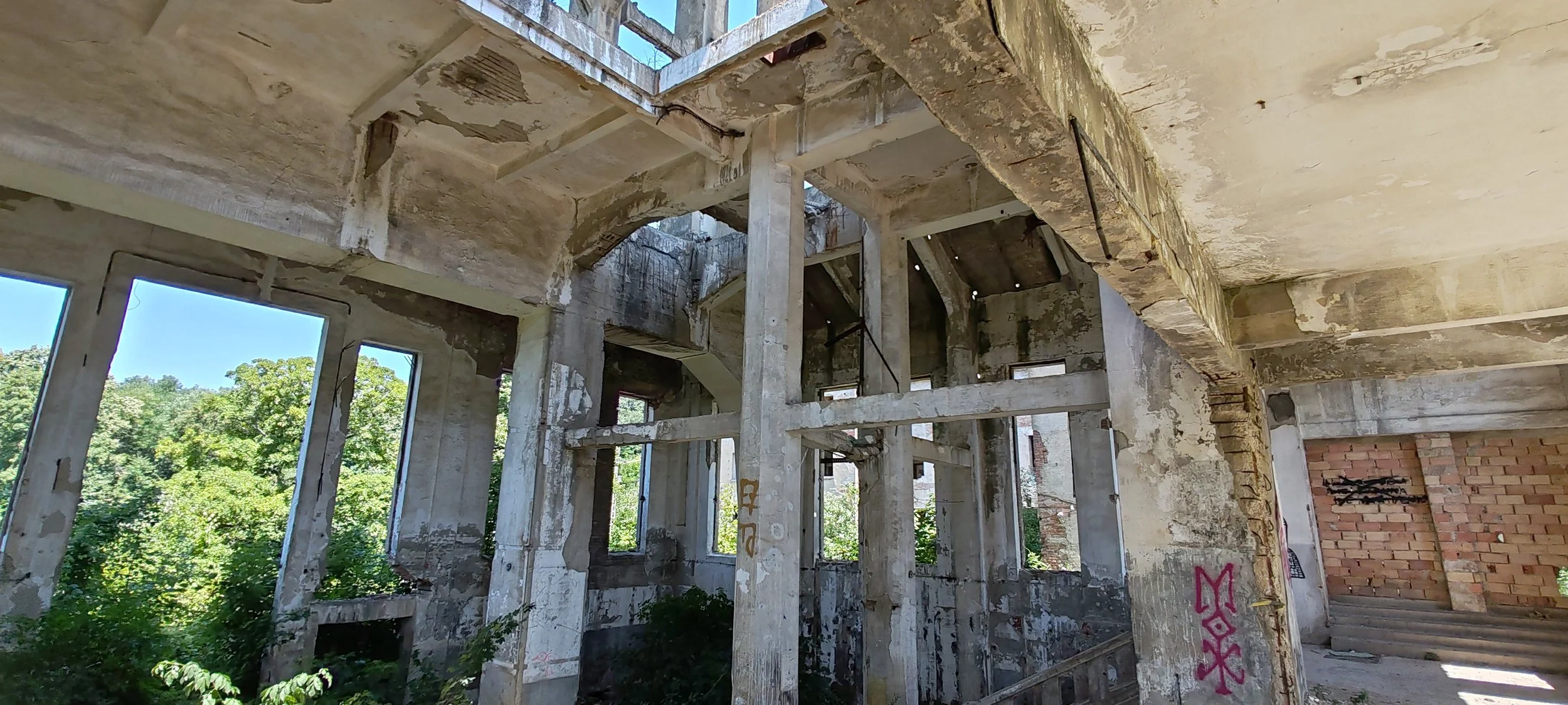 Verlassenes, verfallenes Gebäude mit abblätternden Wänden, großen offenen Fenstern und einer unvollständigen Treppe im Inneren, umgeben von grünen Büschen und Bäumen im Freien.