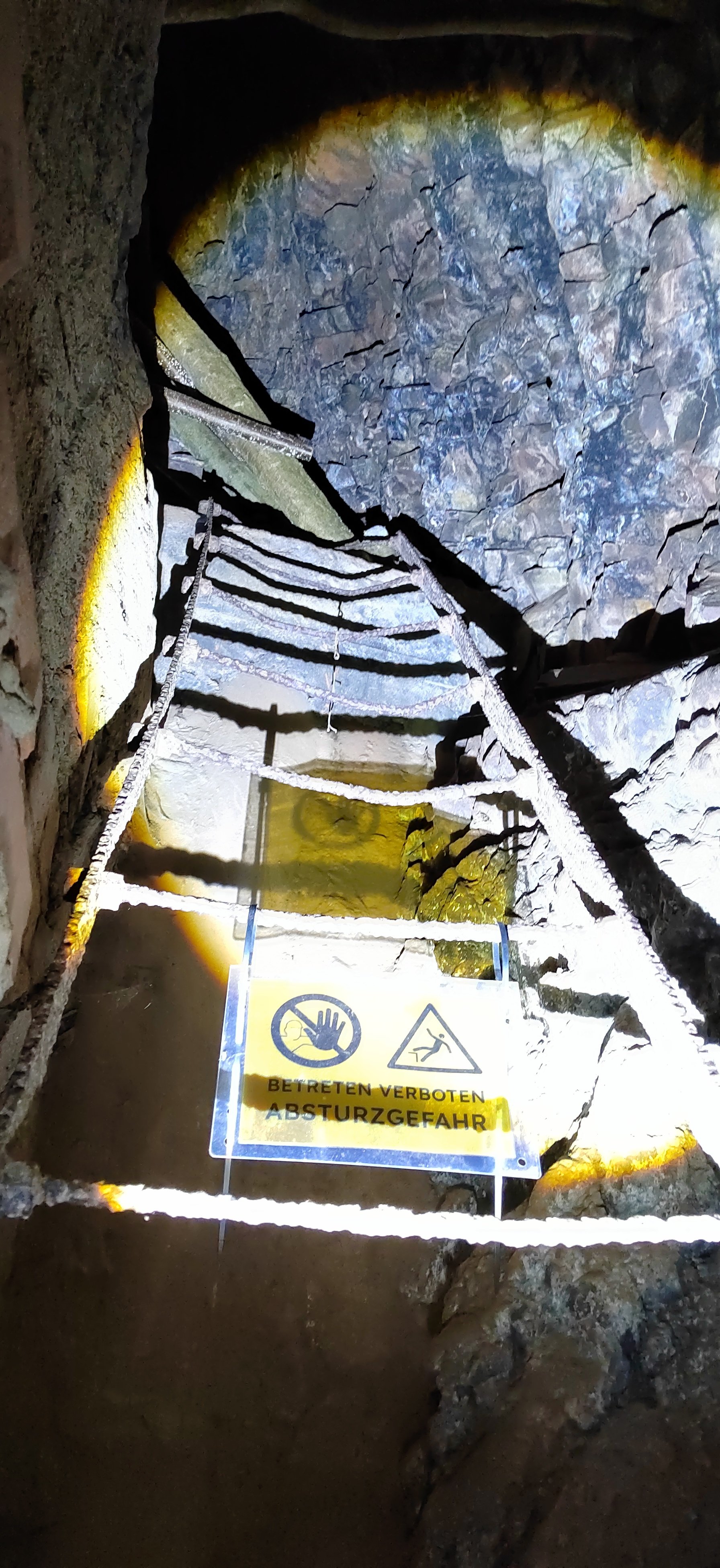 Ein absturzgefährlicher Tunnel mit einer Leiter und einem Warnschild, das das Betreten verbietet. Der Tunnel ist dunkel mit steinartigen Wänden und ist beleuchtet.